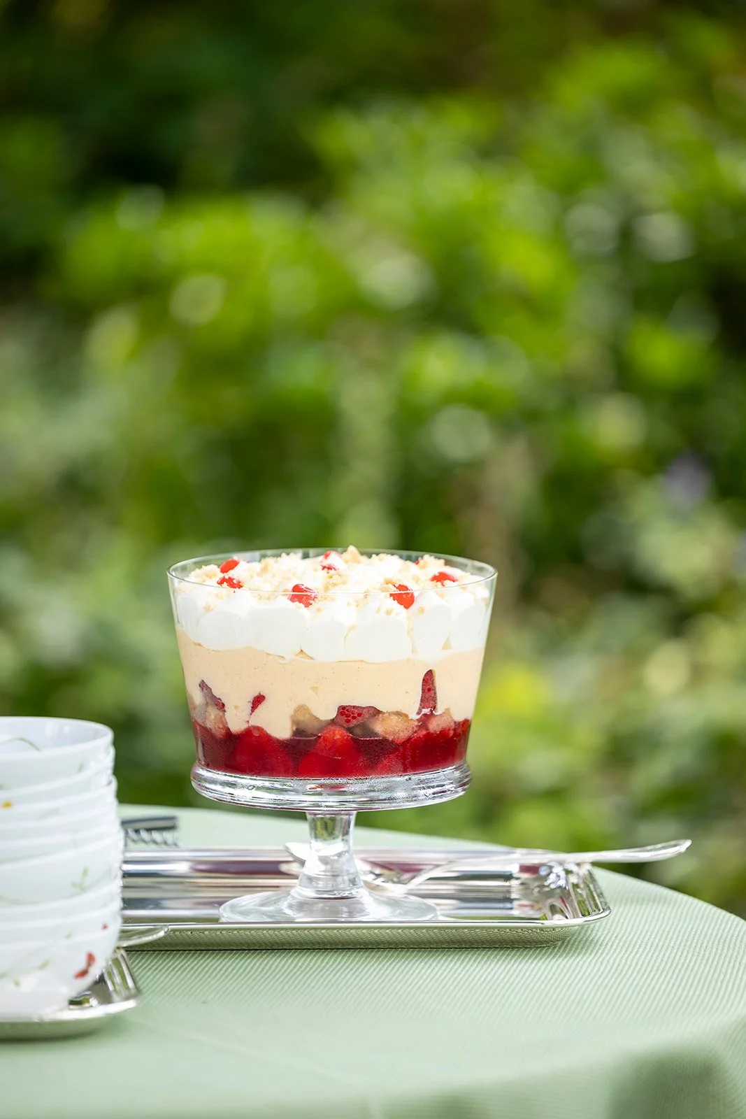 A glass dessert cup filled with layered strawberry shortcake topped with whipped cream and small red fruit pieces, set on a silver tray outdoors with a greenery background.