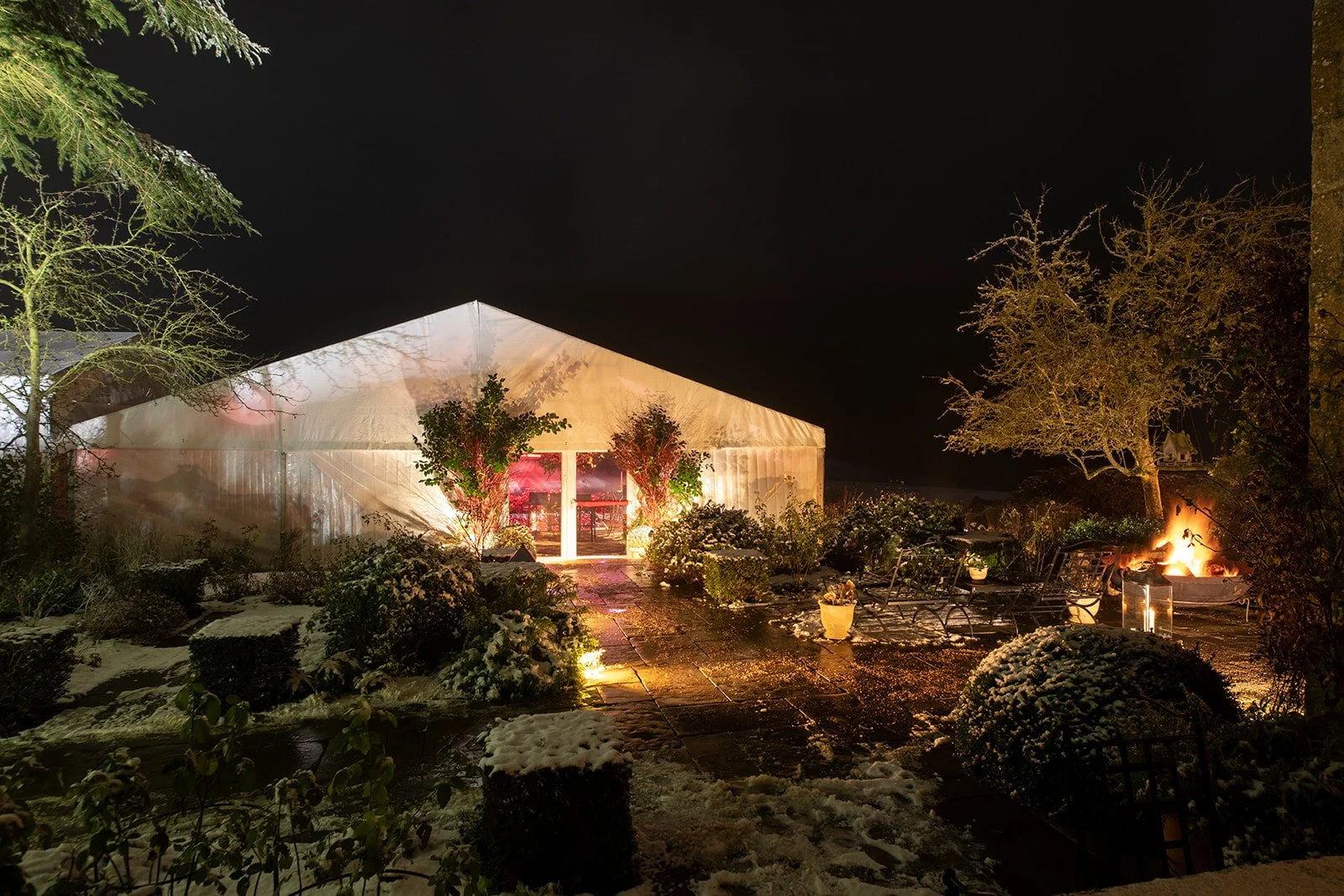 A snowy outdoor garden scene at night with a lit tent, potted plants, trees, and a small fire burning on the right side.