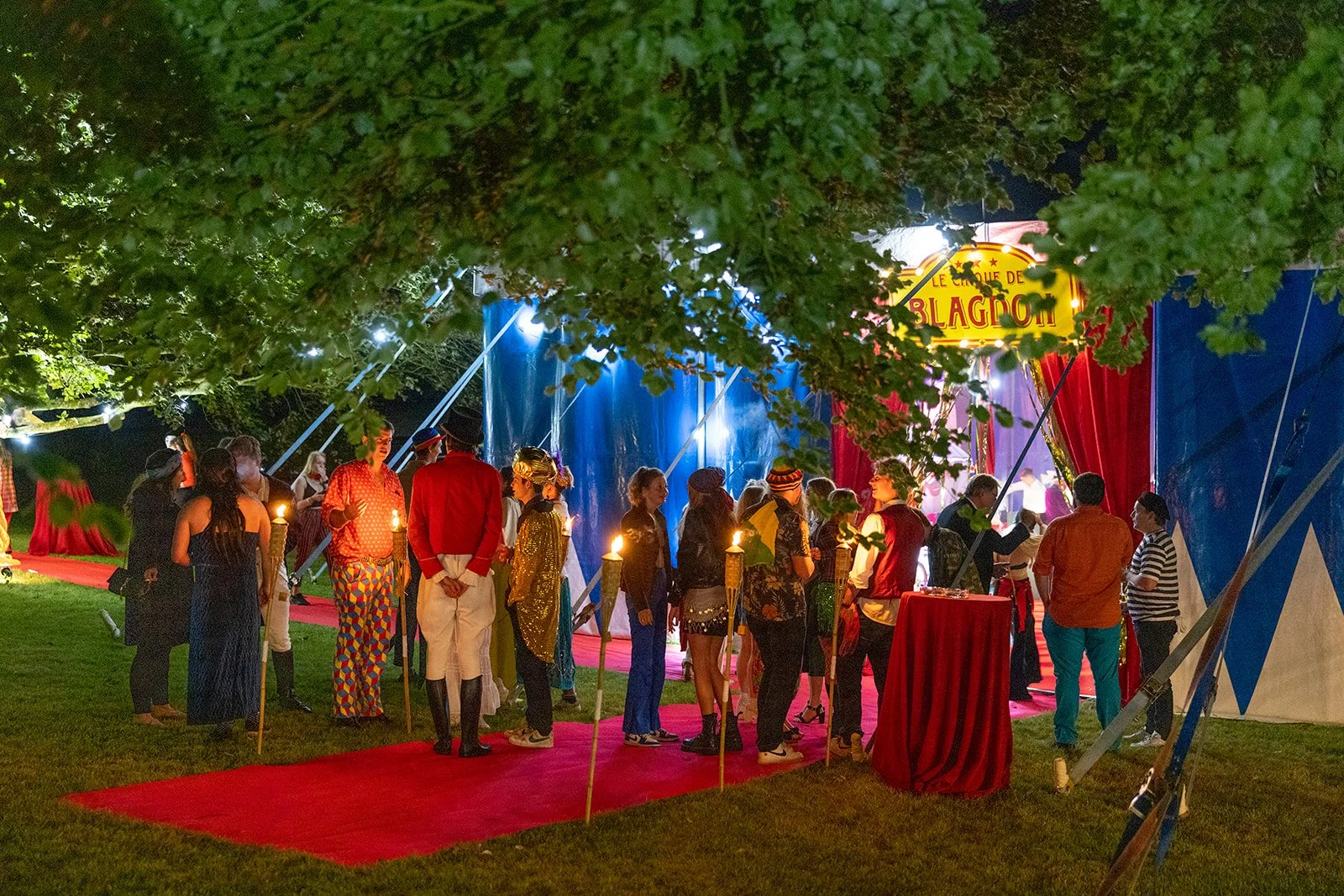 People dressed in colorful costumes and vintage attire gather outside a circus tent with a sign that reads 'Le Cirque de Blagouf'. The scene is illuminated with soft lighting, and a red carpet leads to the entrance, surrounded by green trees at night
