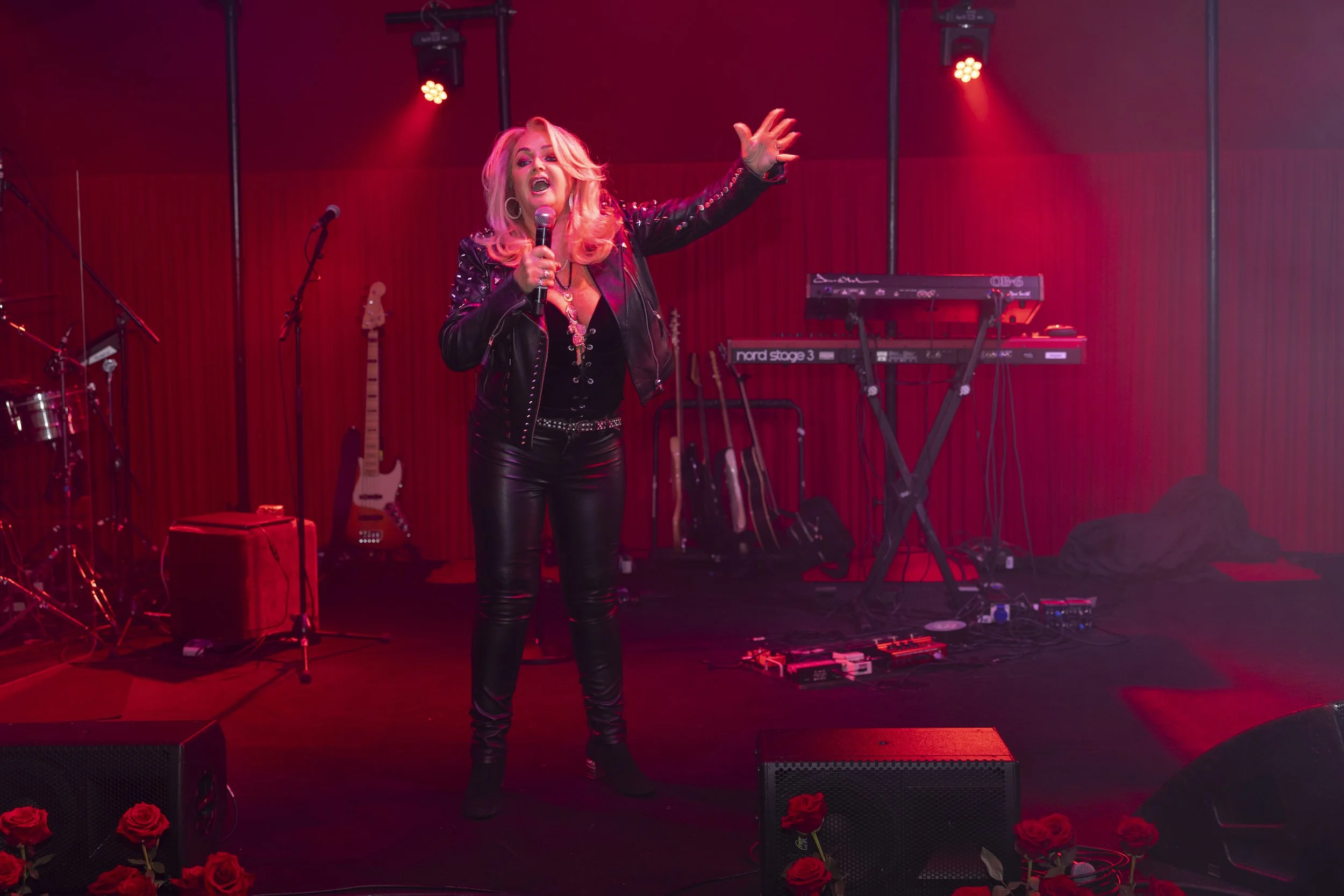 A woman singing on stage holding a microphone, surrounded by musical instruments and equipment, with red lighting and roses at the front of the stage.