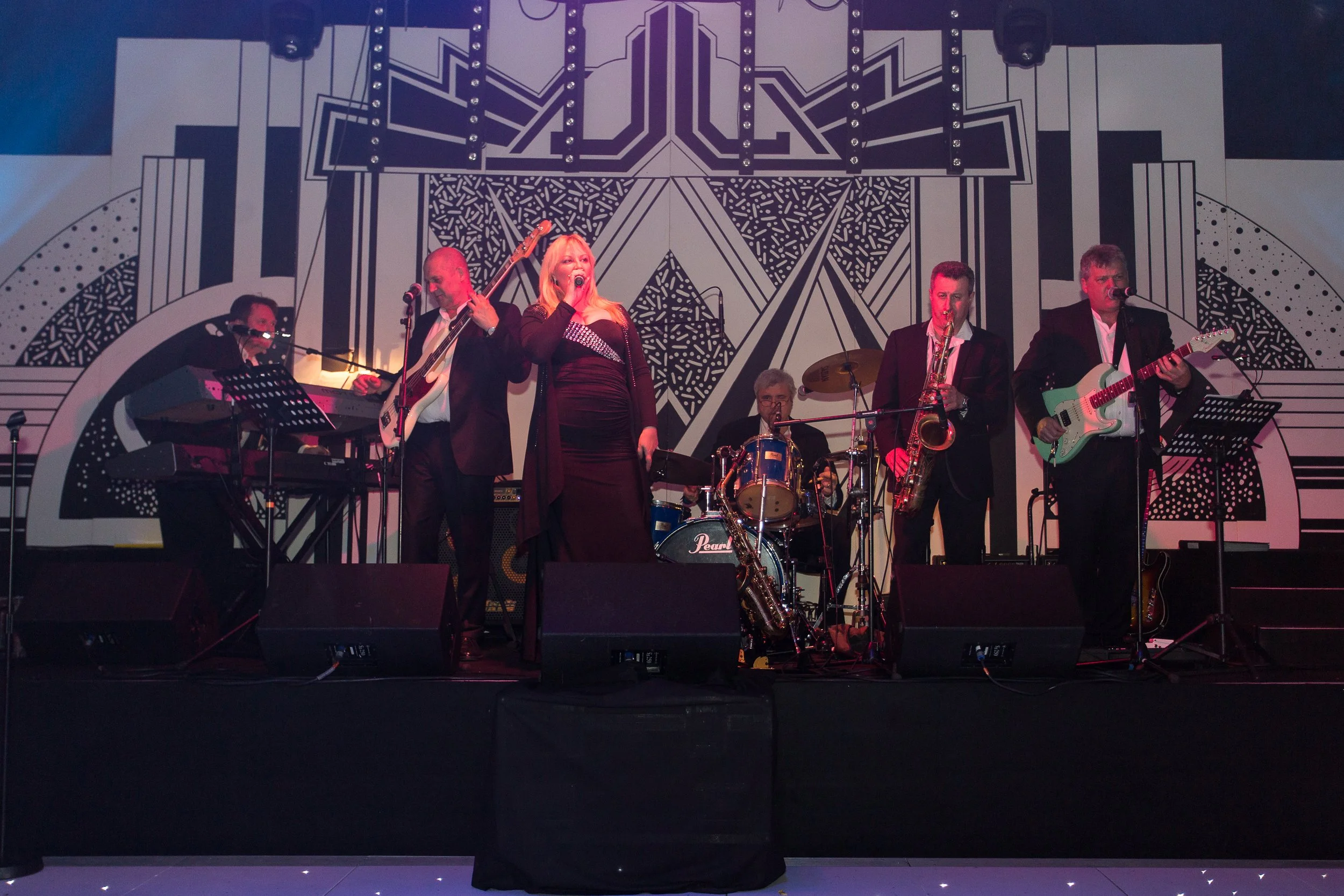 A live band performing on stage with five musicians: a keyboardist, guitarist, vocalist, saxophonist, and drummer. The background features a black and white geometric art piece.