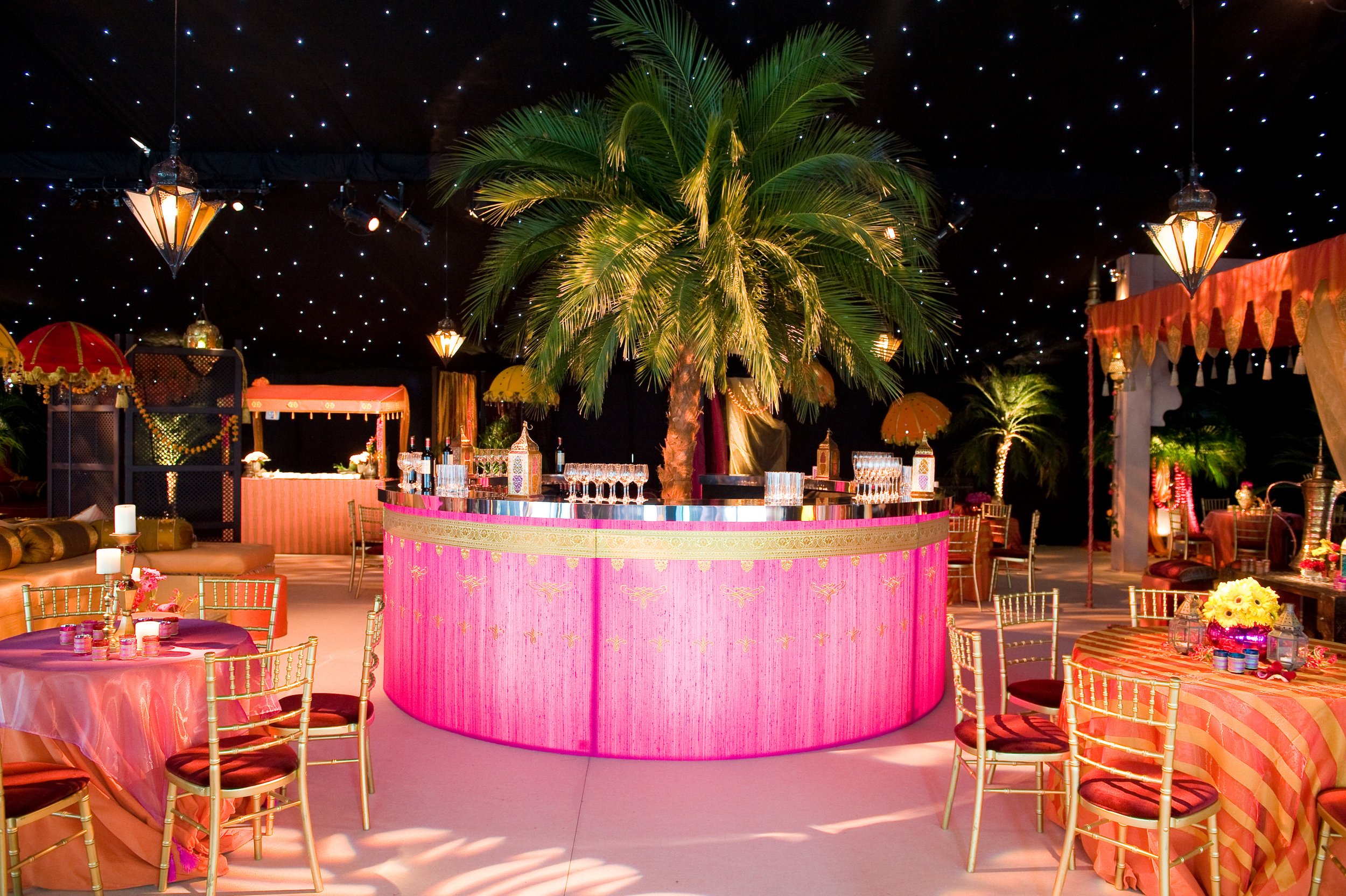 Indoor event space decorated with pink and gold accents, featuring a large palm tree in the center under a starry ceiling. Tables with pink tablecloths and gold chairs surround the space, with candles, flower arrangements, and ornate lanterns as cent