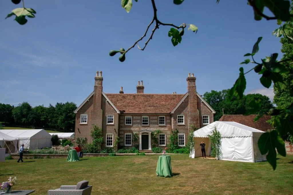 Large historic brick house with a red tile roof, surrounded by a spacious lawn, white tents, and small tables for outdoor event. Greeted by blue sky and green trees.