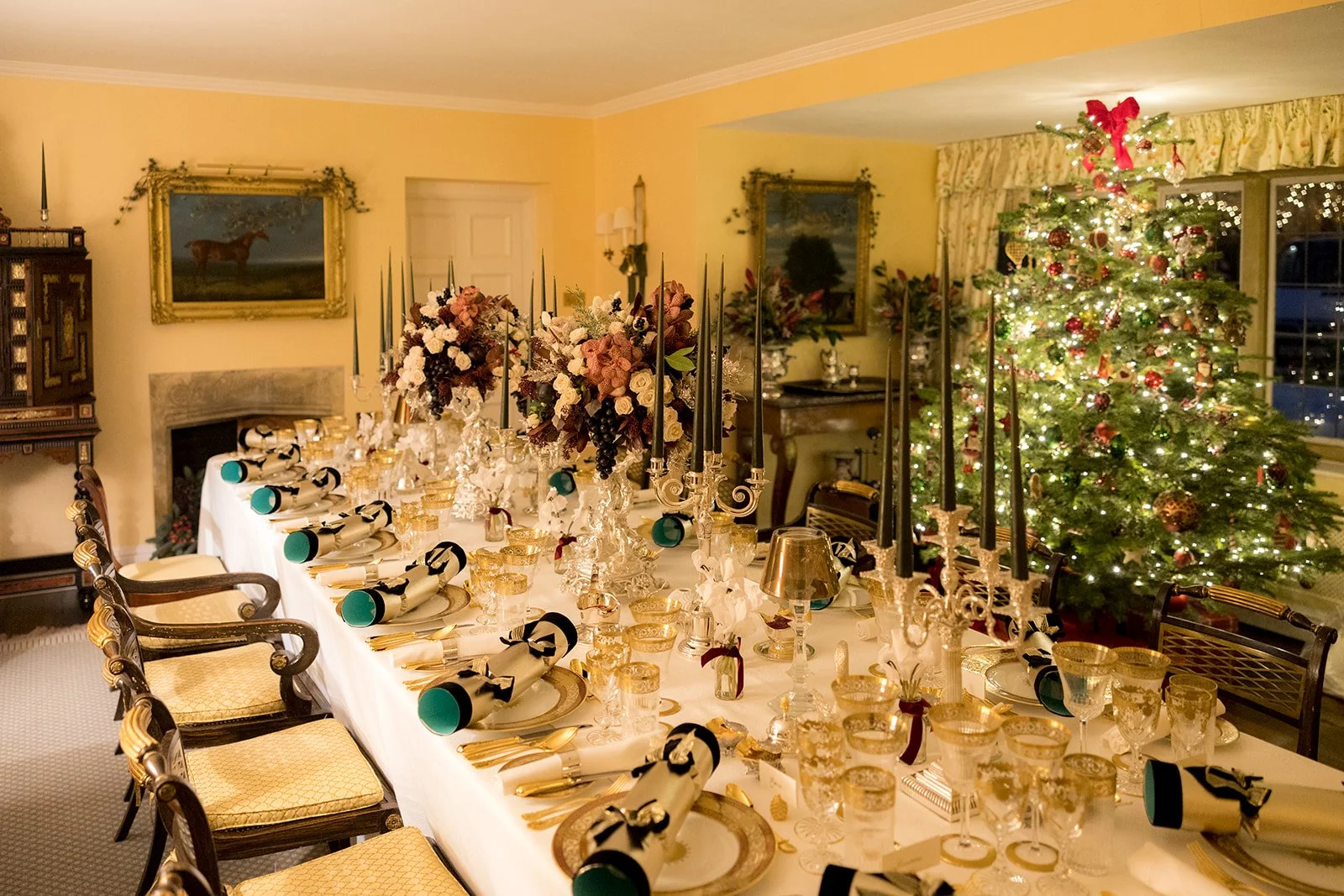 Decorated dining table set for a Christmas celebration with floral centerpieces, candles, glasses, and tableware, with a decorated Christmas tree with lights and ornaments in the background.