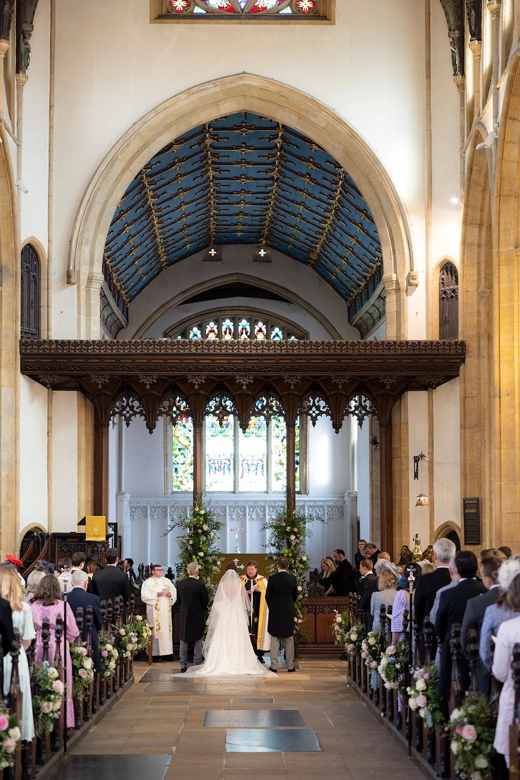 A wedding ceremony inside a church with a vaulted ceiling, stained glass windows, and floral decorations. The bride and groom stand before a priest, surrounded by seated guests.