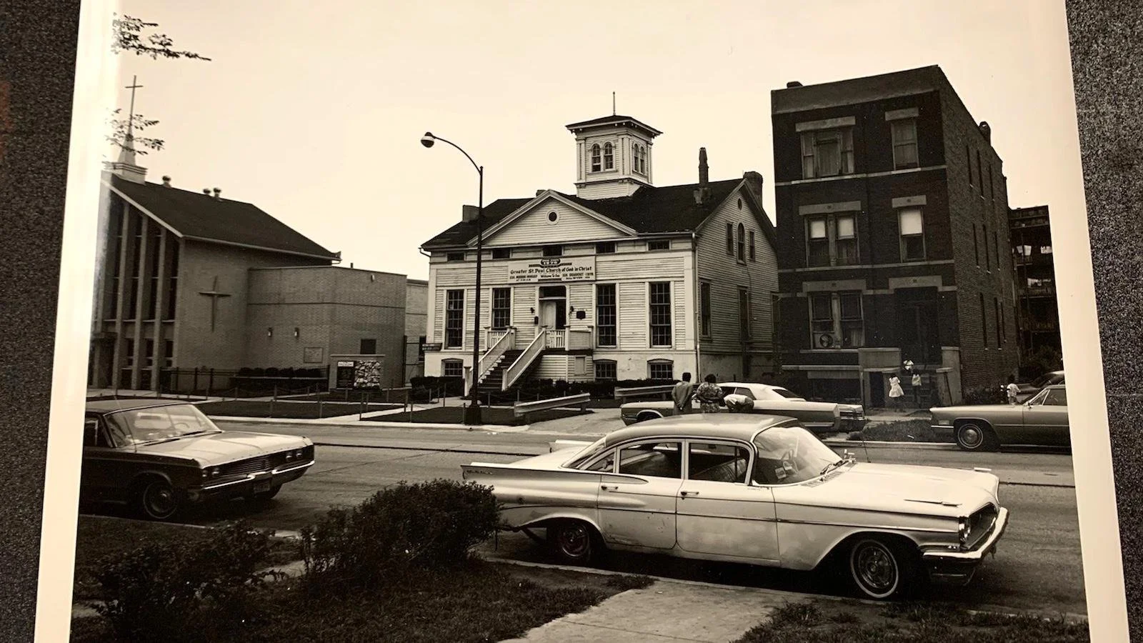 Black and white photo of a church with a small steeple, surrounded by cars and buildings, with a few people walking nearby.