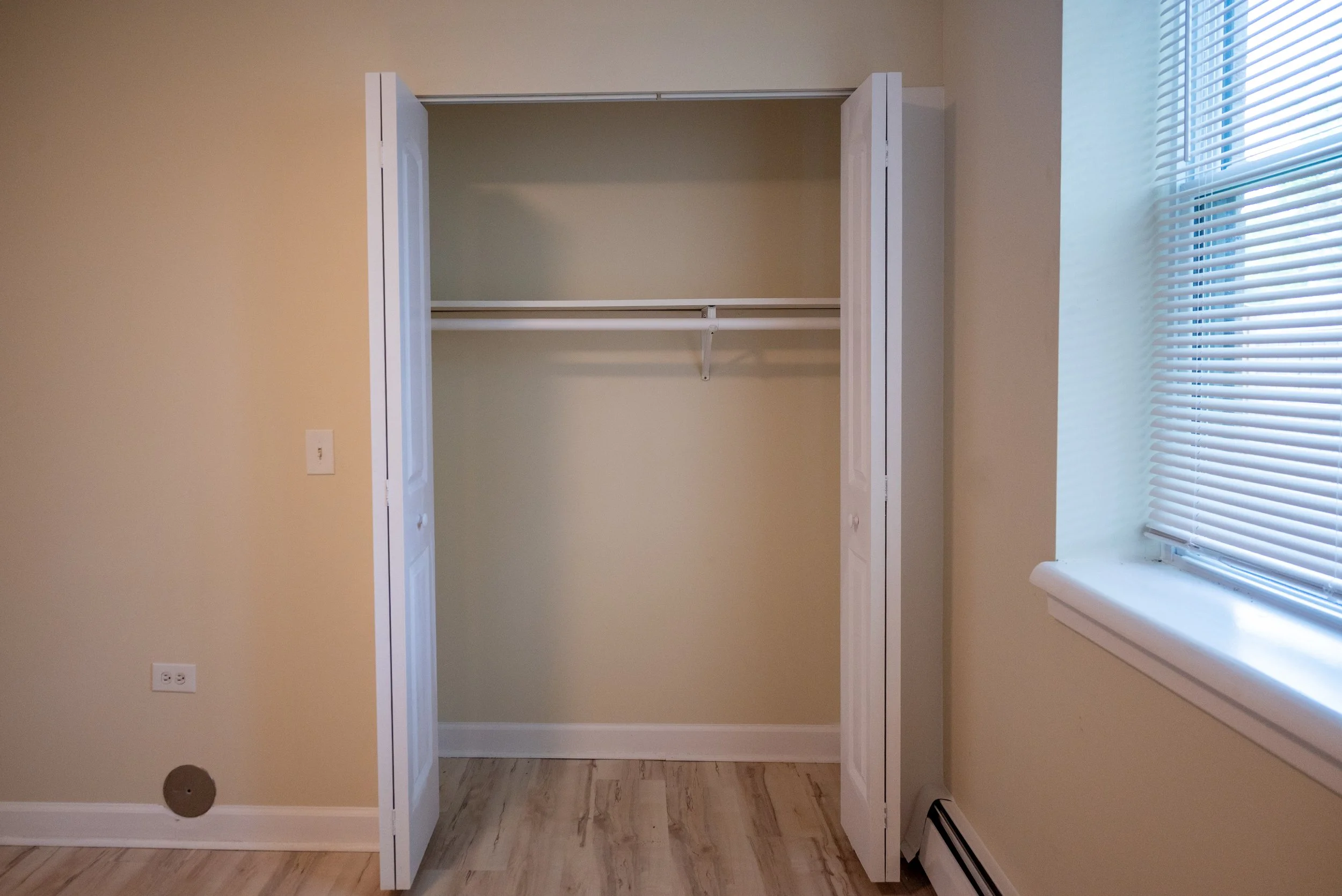 Empty closet with open double doors, a single shelf, and hanging rod inside, next to a window with blinds.
