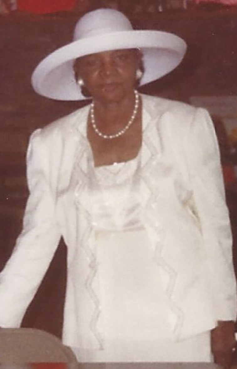 A woman dressed in elegant white clothing, wearing a large white hat, pearl necklace, and earrings, standing outdoors at night.