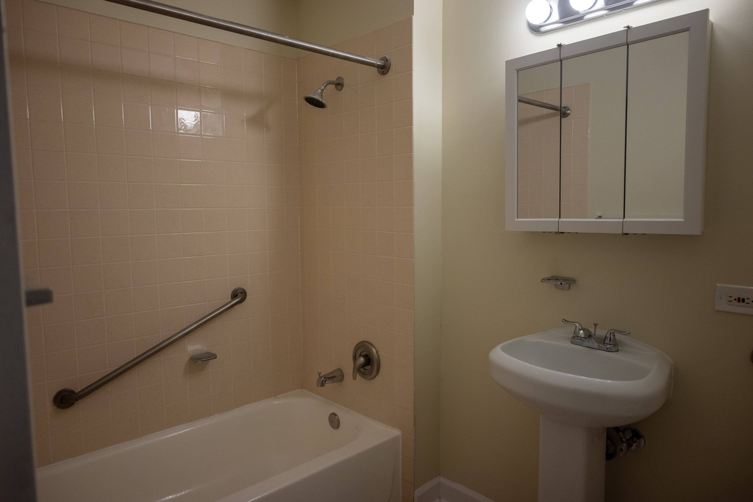 A beige-tiled bathroom with a bathtub, grab bars, a showerhead, a small mirrored cabinet above a sink, and a wall-mounted mirror with lights.