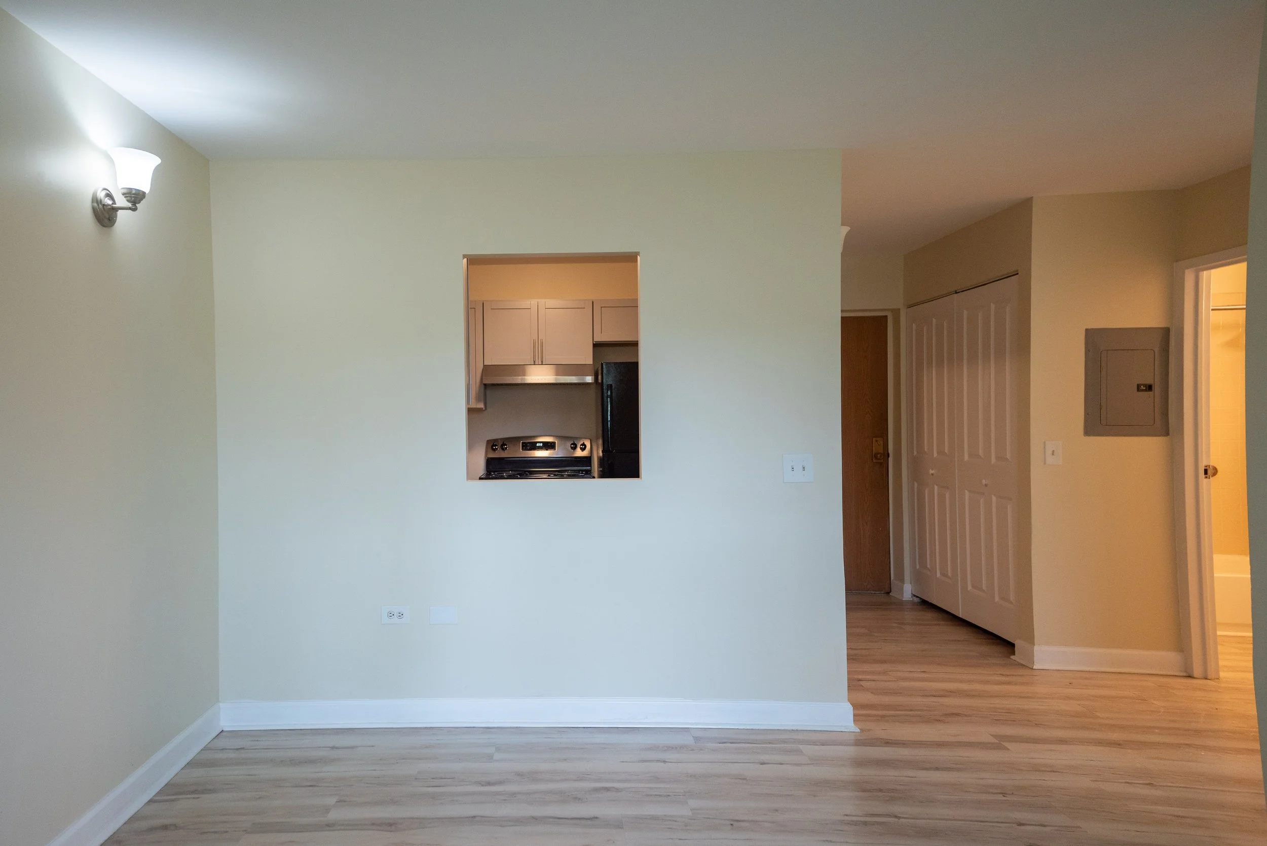 Interior view of an empty living room with light green walls, wood flooring, a wall-mounted light fixture, a small open kitchen area with light-colored cabinets and a stove, and a hallway with a closet and an electrical panel.
