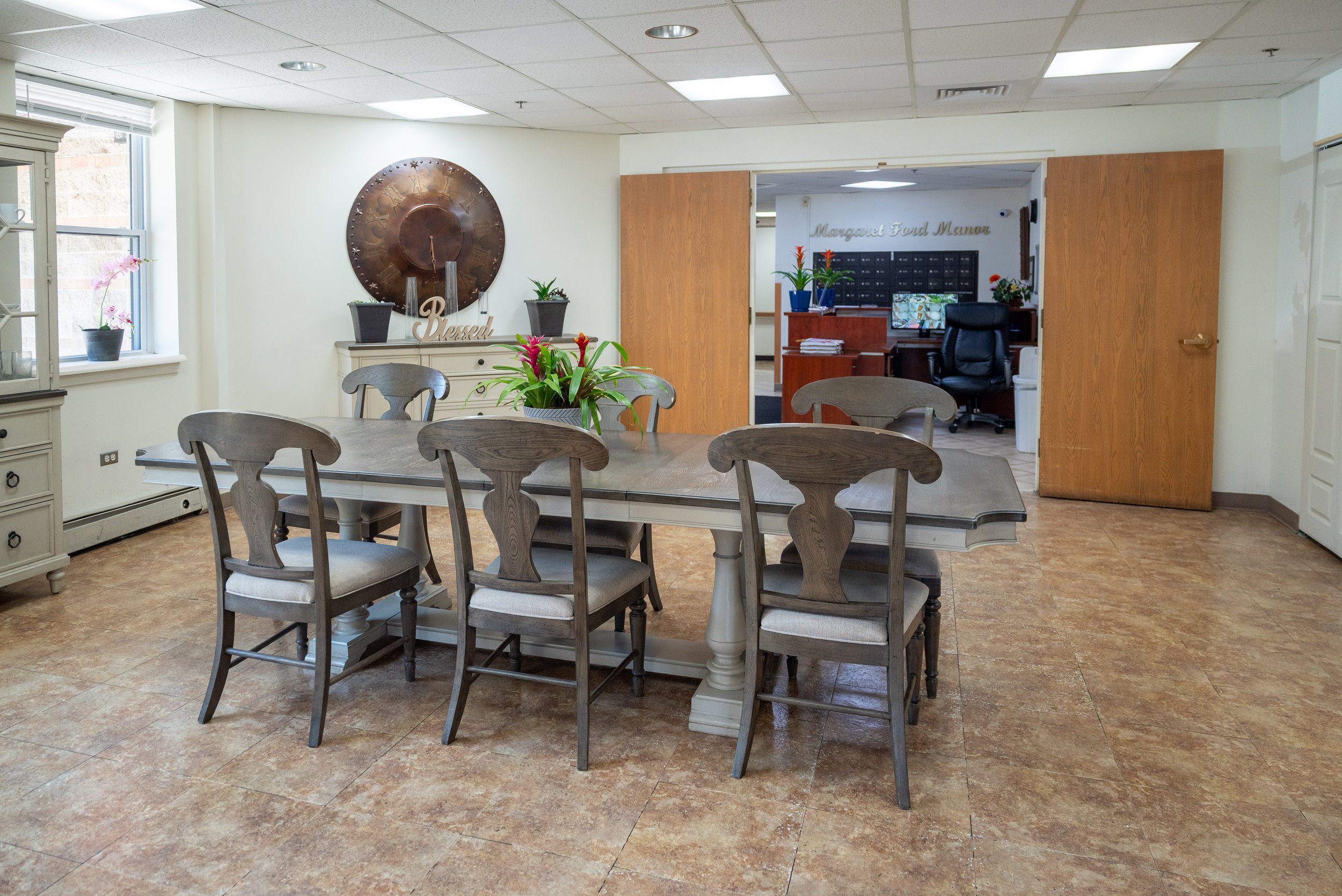 A dining area with a large wooden table, several gray wooden chairs, and decorative plants. In the background, a reception area with mailboxes and a sign reading 'Margaret Ford Manor' is visible through an open doorway.