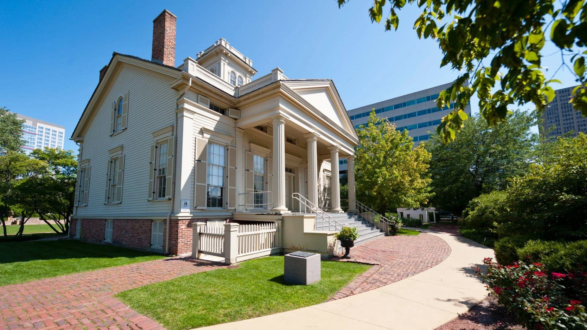 A historic white house with tall columns, a brick foundation, a staircase, and large windows, surrounded by trees and a brick walkway, with modern buildings in the background.