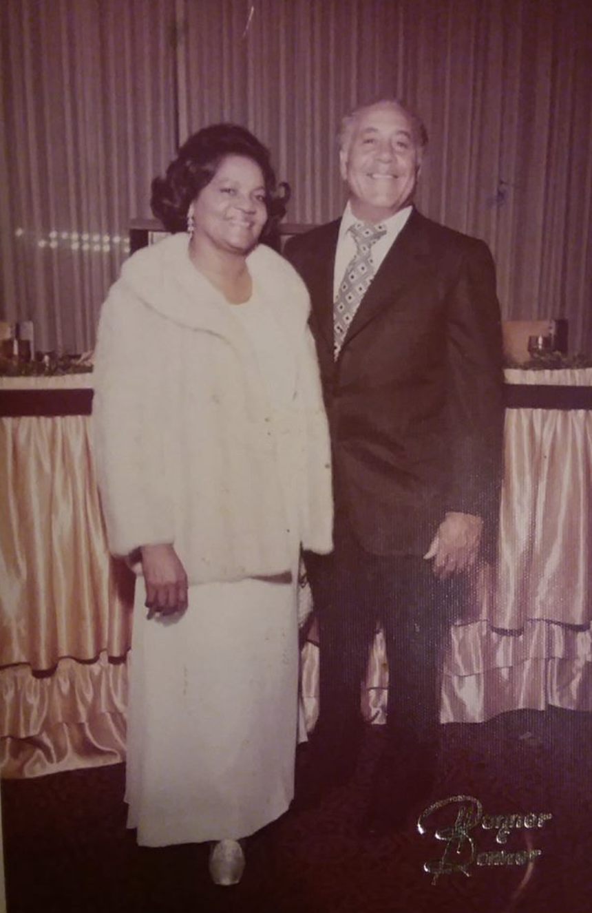 A woman and a man standing side by side in a room with wood-paneled walls and a table with a tablecloth behind them. Both are smiling.