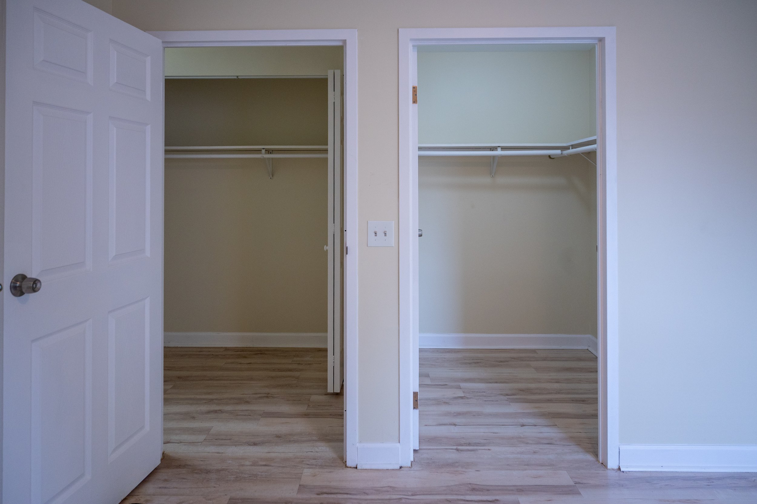 Empty closets with white doors, one open and one partially open, inside a room with light-colored walls and wooden flooring.