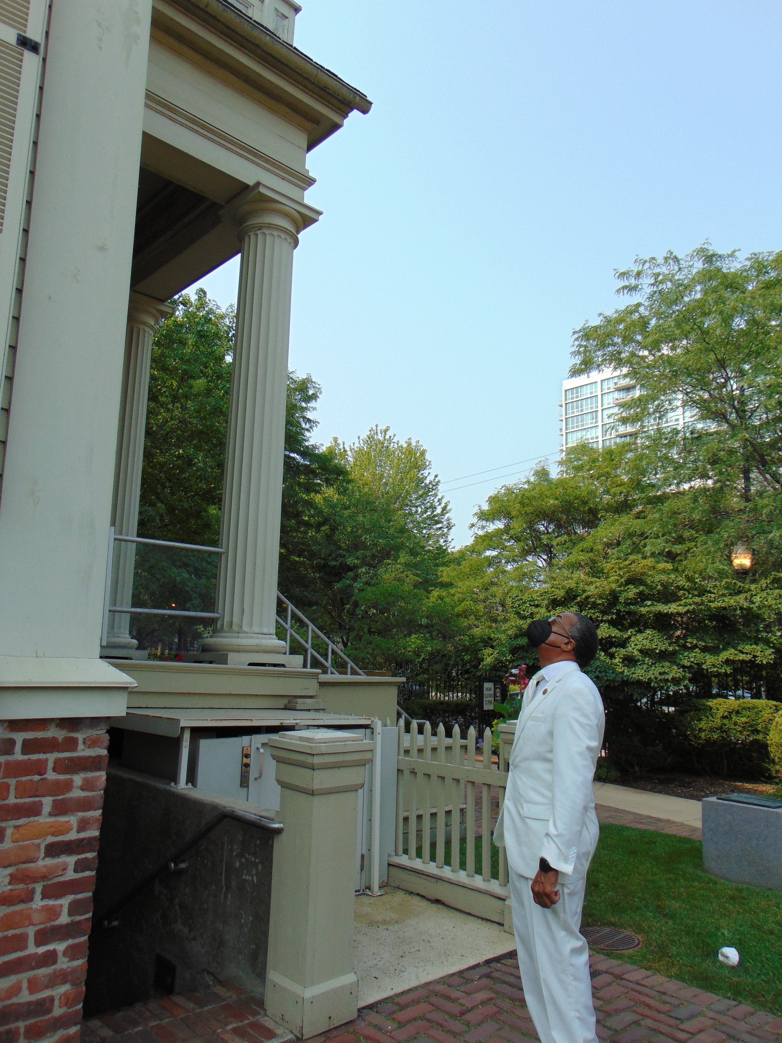 A man in a white suit and black mask standing outside near a building with columns and a staircase, with trees and an apartment building in the background.