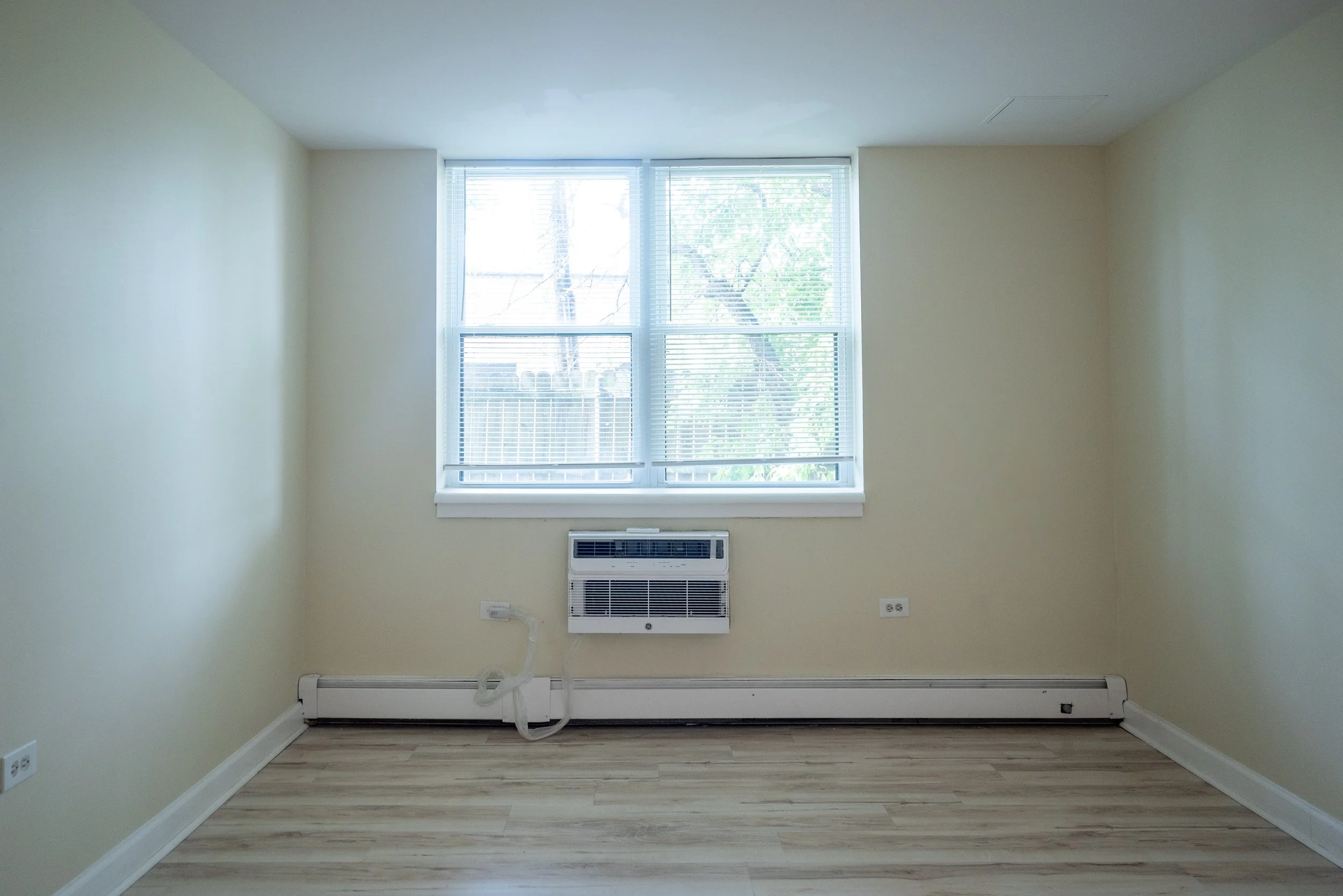 Empty room with beige walls, wooden floor, window with blinds, and wall heater underneath the window.