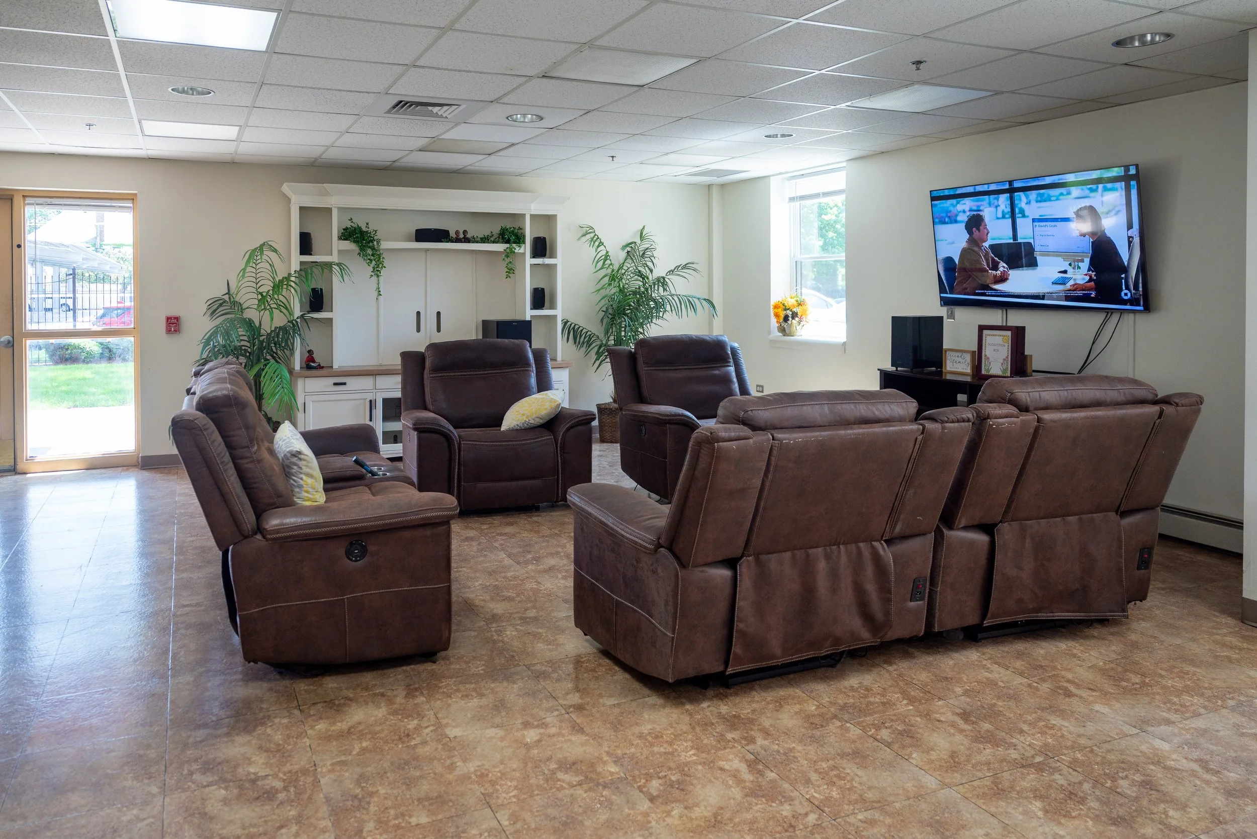 A waiting room with brown leather chairs, a white built-in shelf, indoor plants, a window with sunlight, and a large flat-screen TV.