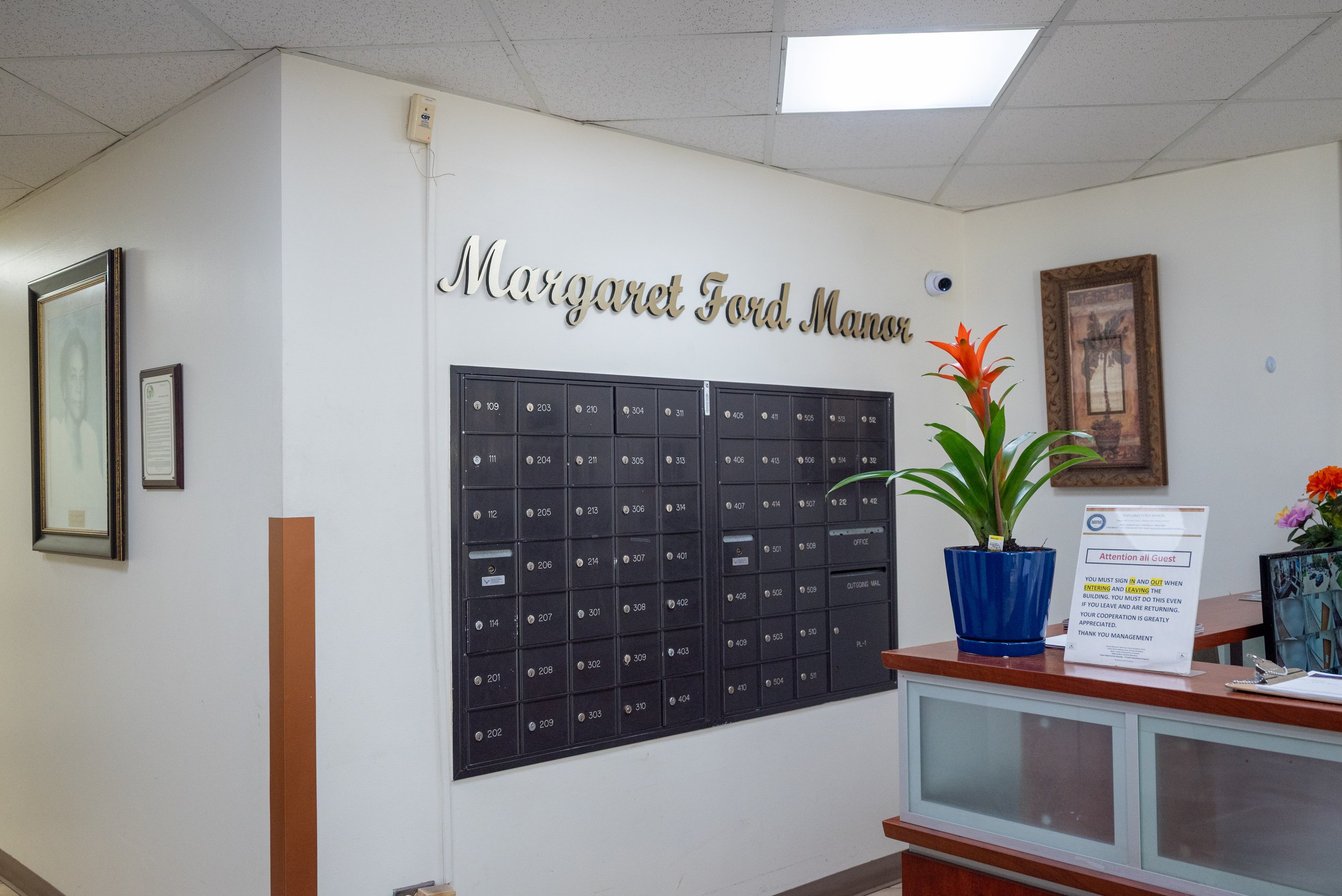 Interior of a building with a wall of black mailboxes, a sign that reads Margaret Ford Manor, framed picture on the wall, a large blue flowerpot with a red flower, and informational signs on a desk.