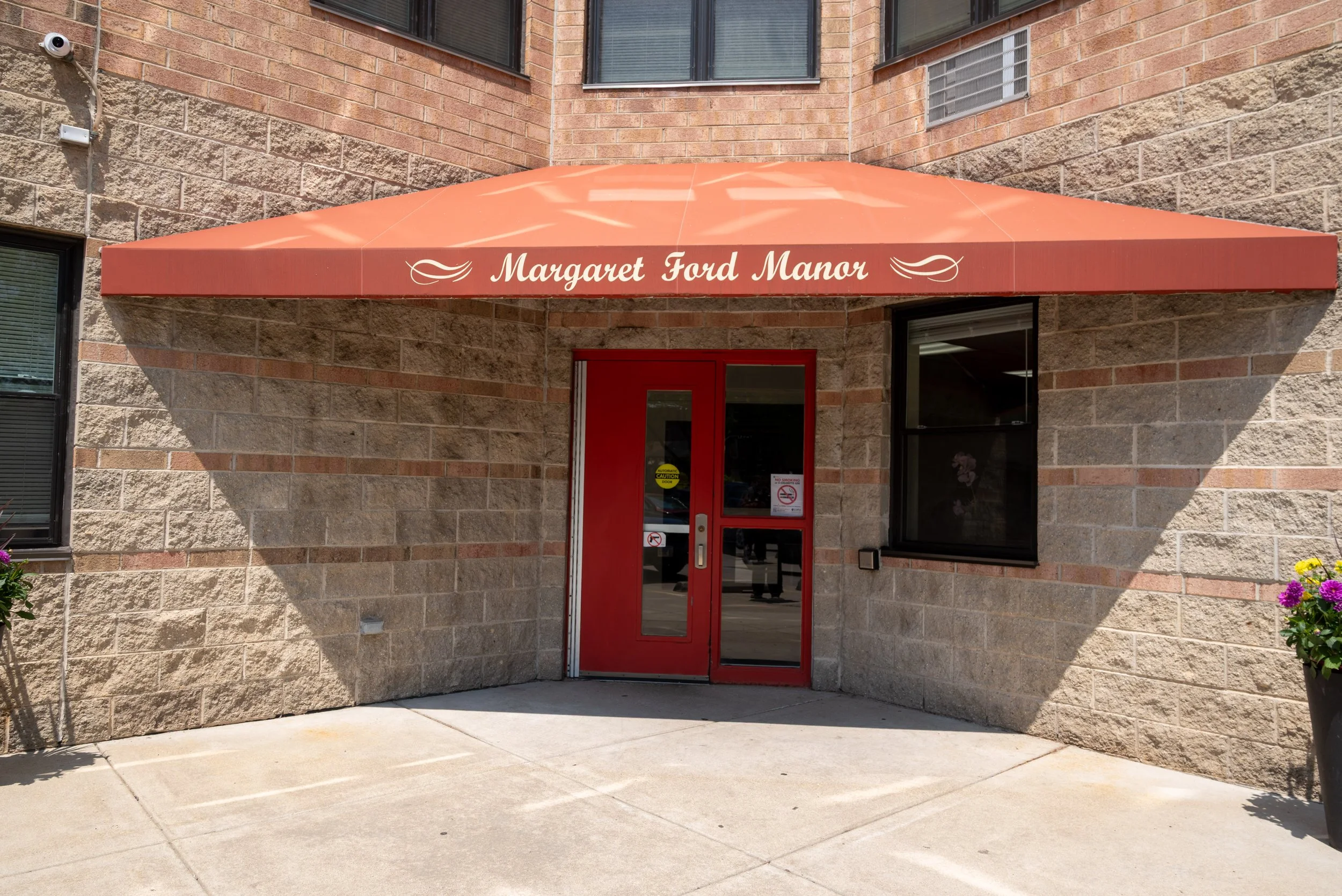 Red door and window with a red awning that reads 'Margaret Ford Manor' on a brick building. There are flowers in pots nearby.