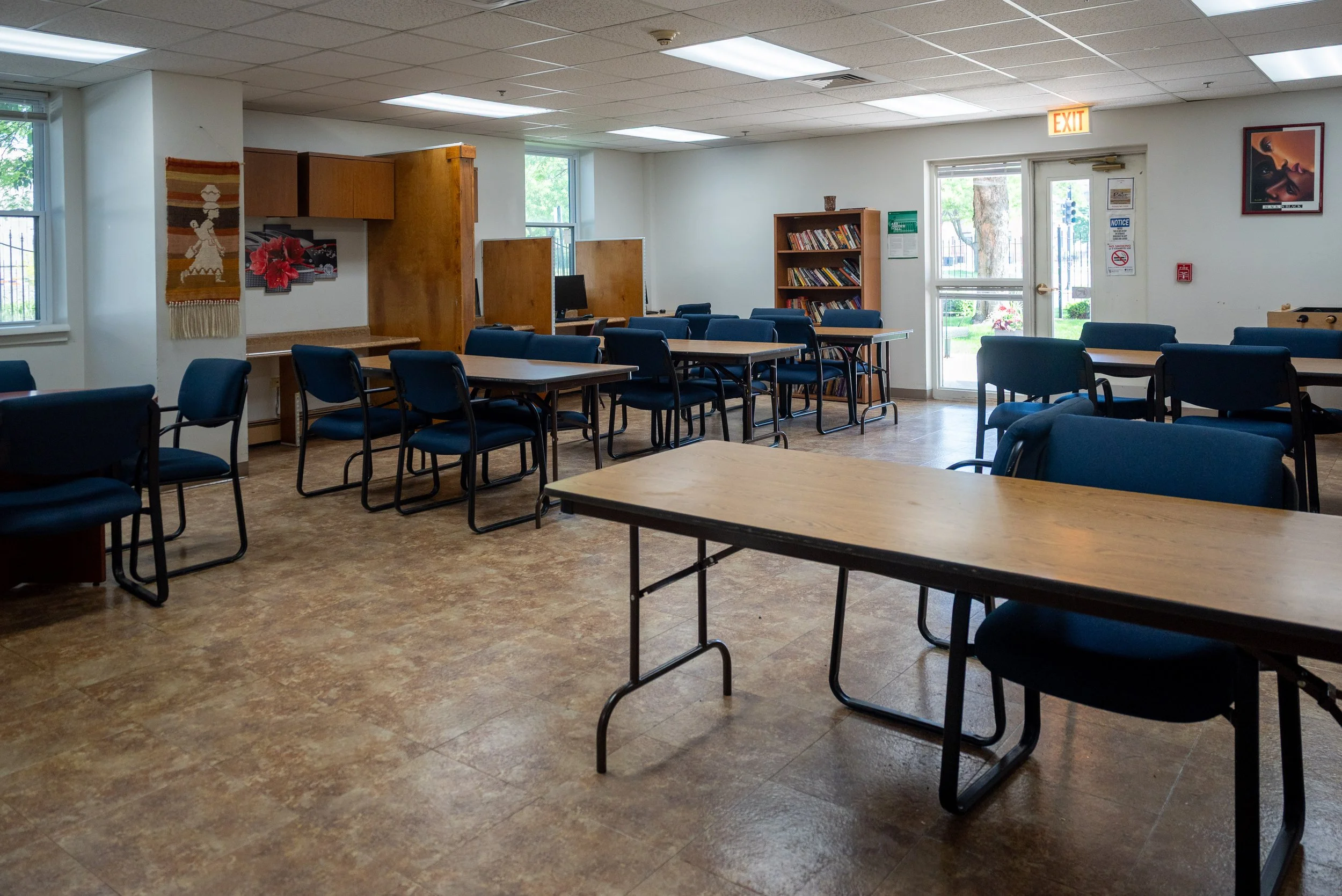 Empty community center or classroom with blue chairs, wooden tables, bookshelves, windows, and colorful wall art.