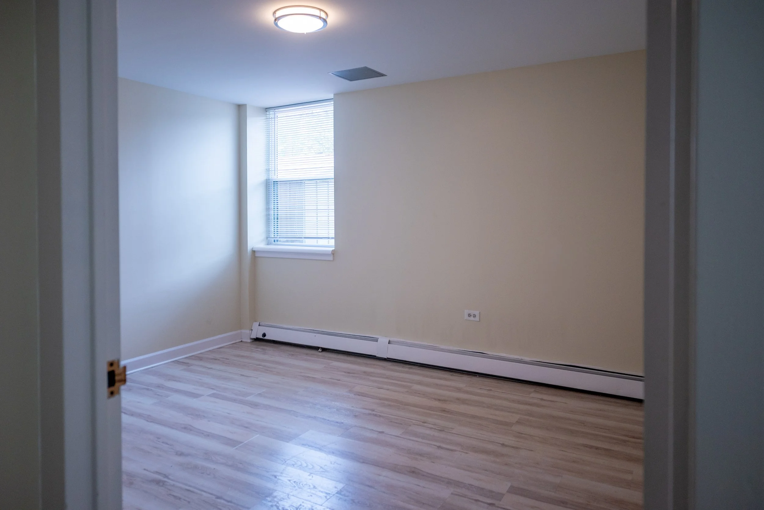 Empty room with a window that has venetian blinds, a ceiling light fixture, baseboard heater, and hardwood flooring.