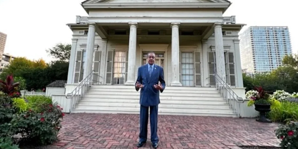 A man in a blue suit standing on a brick path in front of a large white historic house with columns, stairs, and gardens.