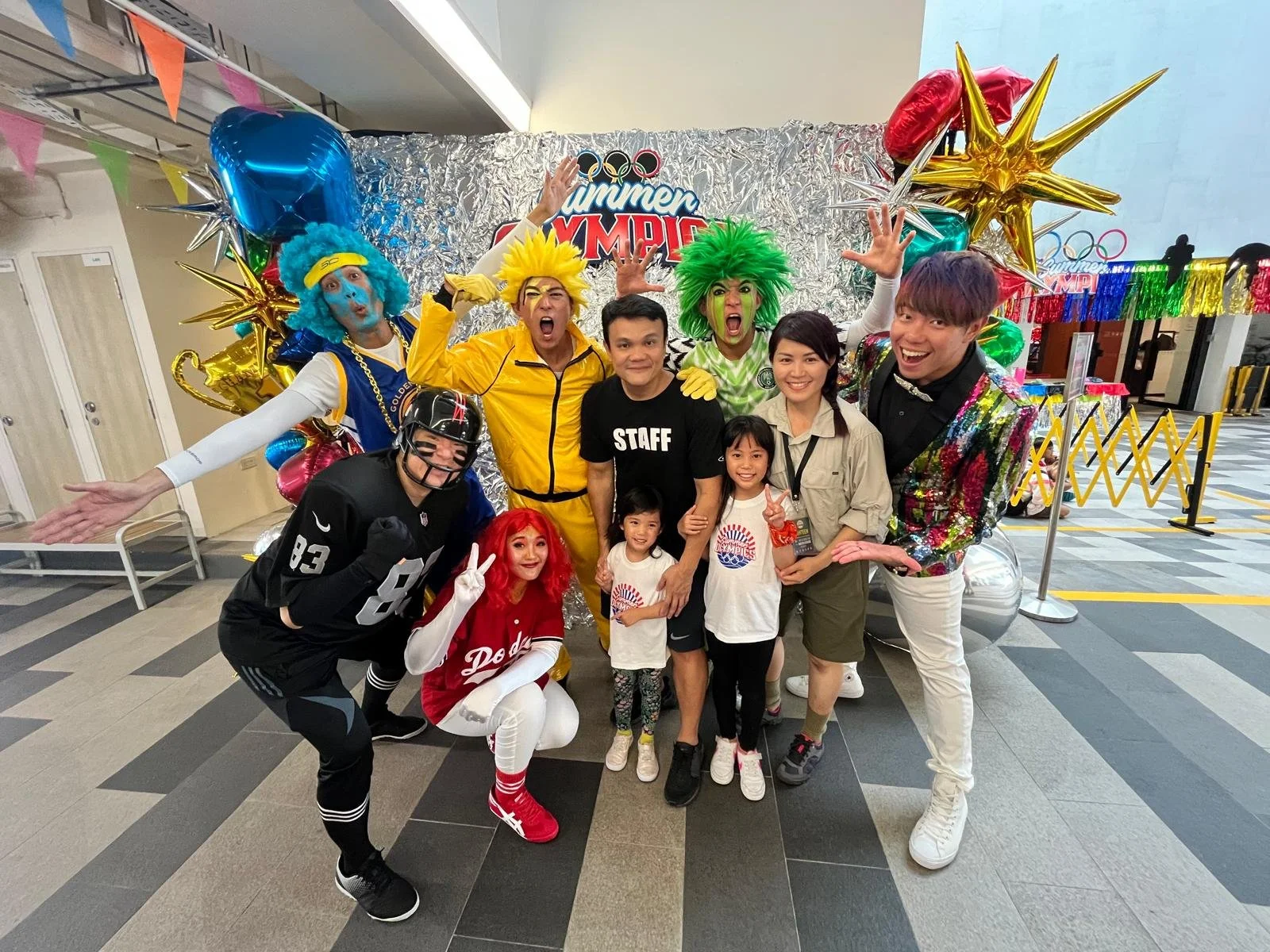A group of people, including three children, posing with colorful entertainers dressed as clowns and athletes at a summer event with balloons and a shiny backdrop.
