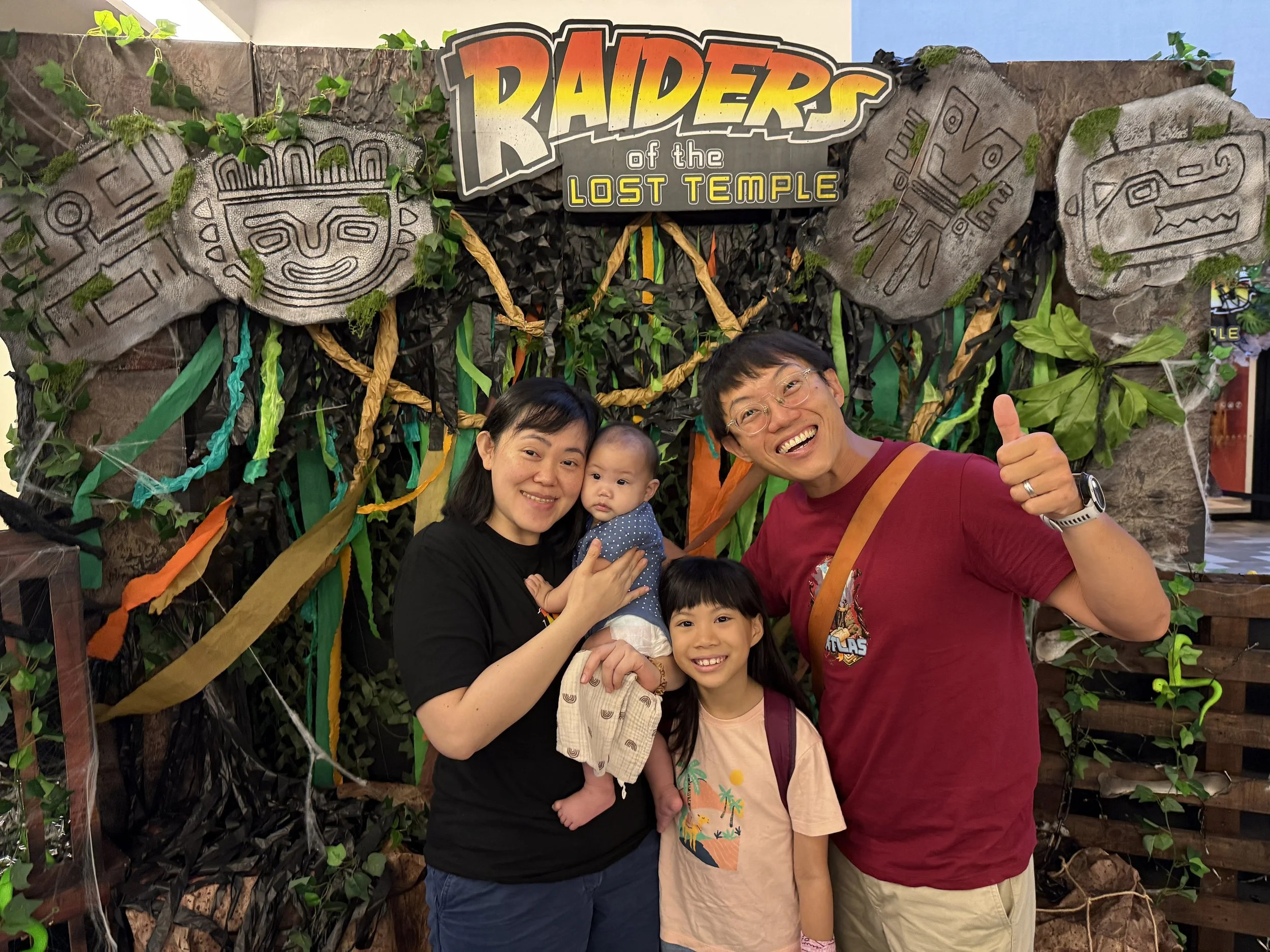A smiling family of four, including two women and two children, posing in front of a decorative jungle-themed backdrop with a sign that reads 'Raiders of the Lost Temple,' surrounded by artificial plants and carved stone designs.