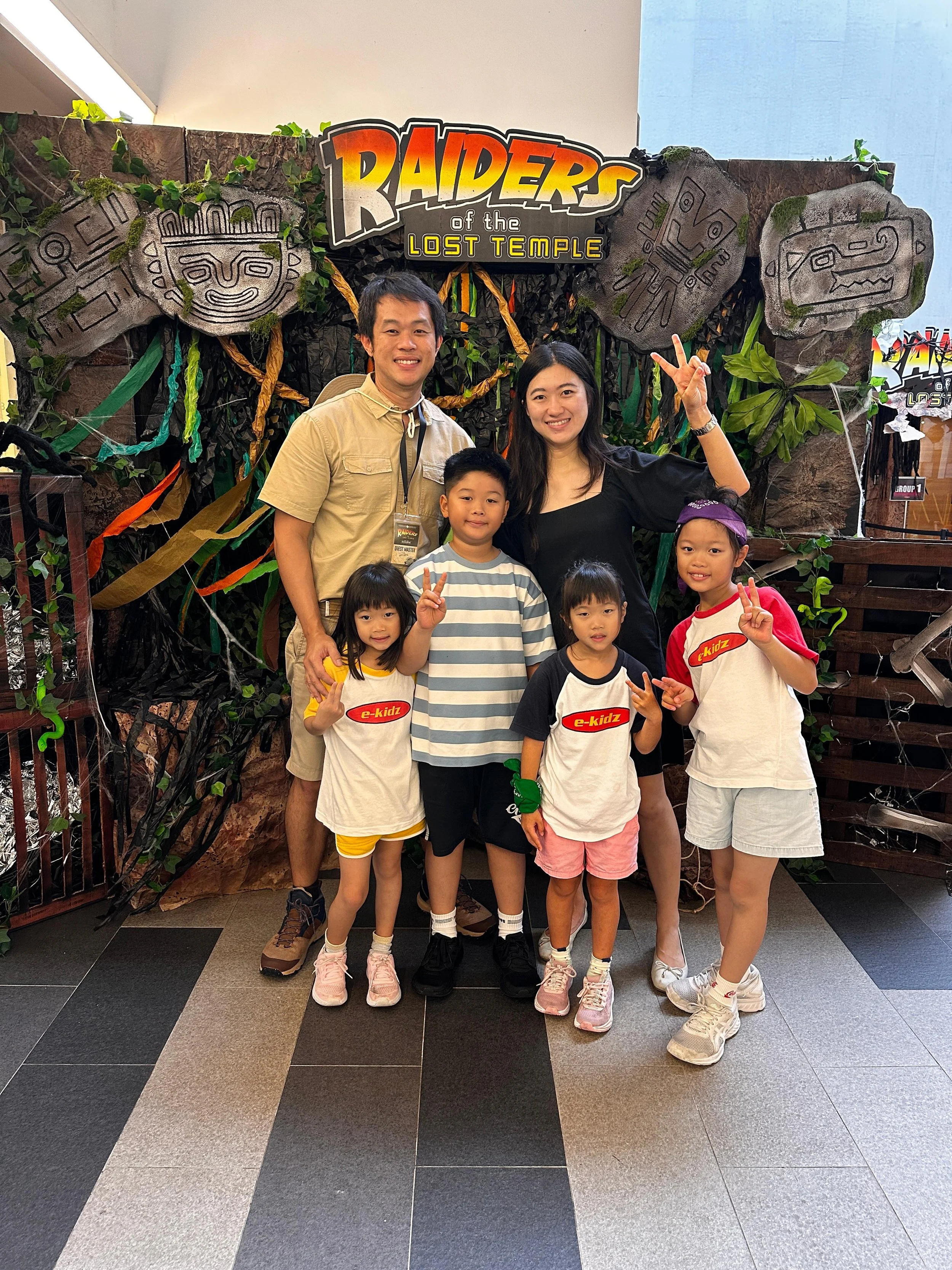 Group of six children and two adults standing in front of a Jungle-themed backdrop with a sign that says "RAIDERS of the LOST TEMPLE." The children are wearing casual clothes, and some are making peace signs. The adults are smiling, one making a peace sign.
