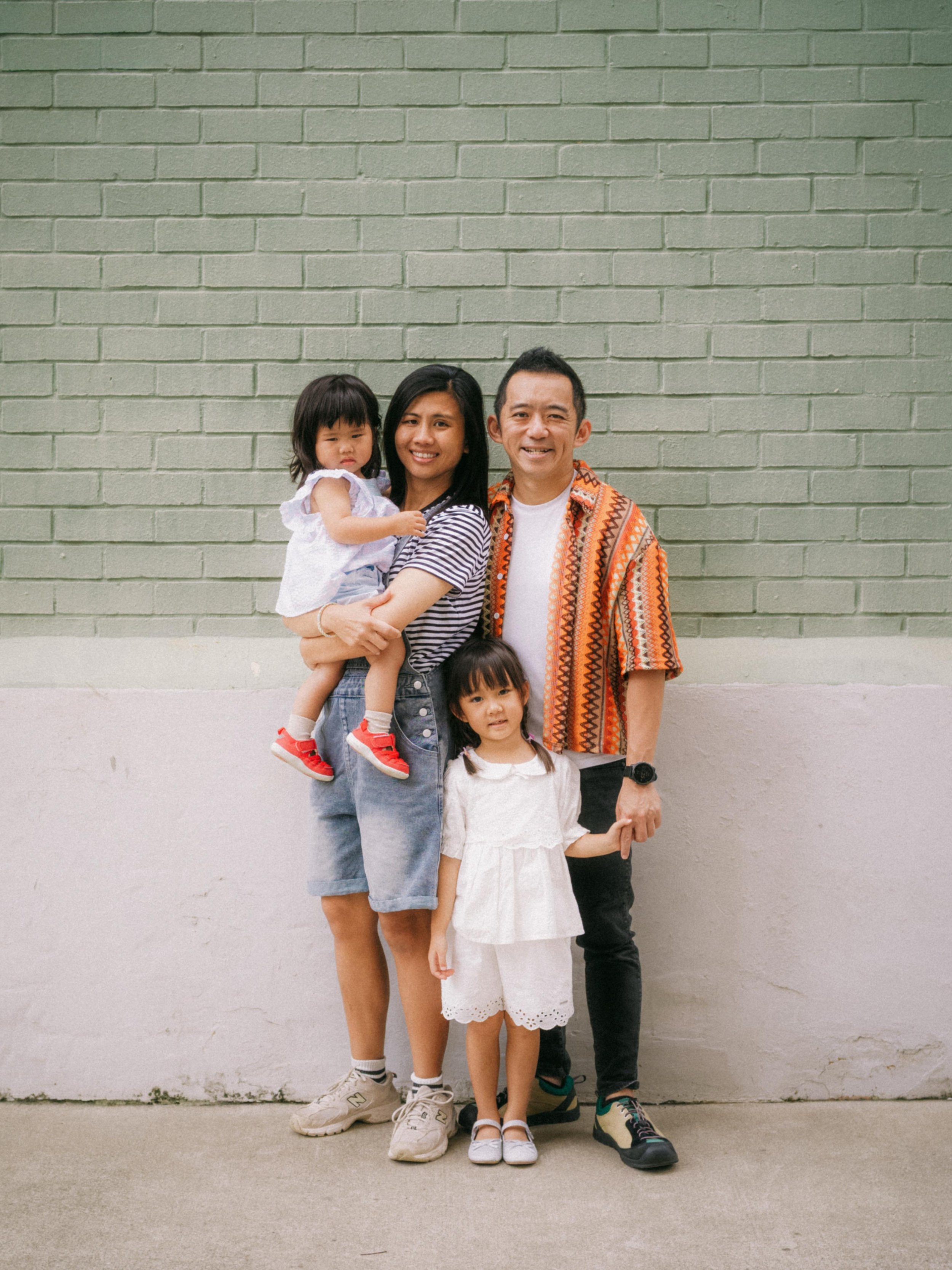 A family of five standing in front of a green brick wall, including two adults and three children. One woman is holding a young girl, and an older girl stands in front of the adults, with a man standing beside them.