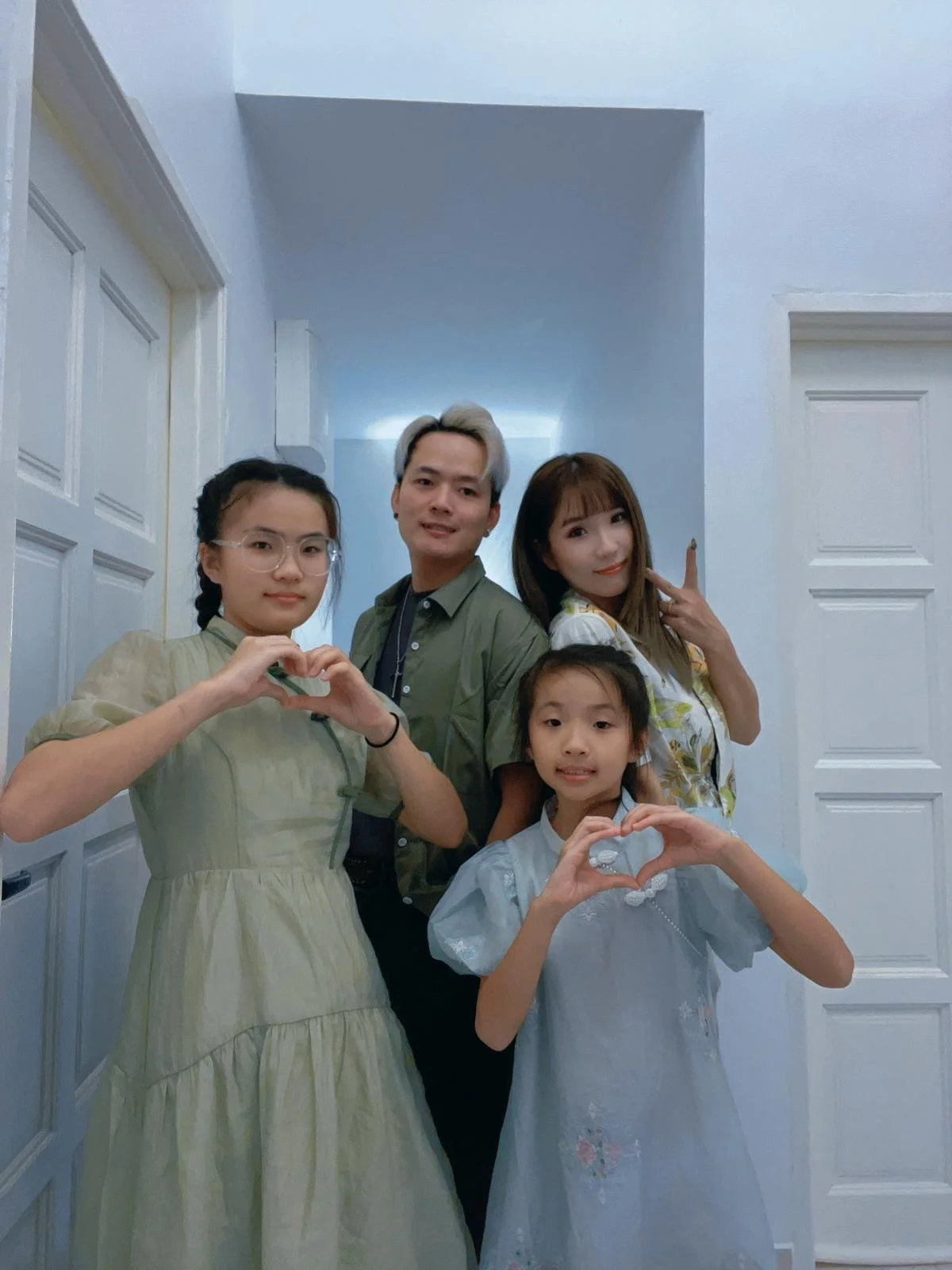 Group of five people standing together indoors, forming heart shapes with their hands, smiling, with a light blue wall and white doors in the background.
