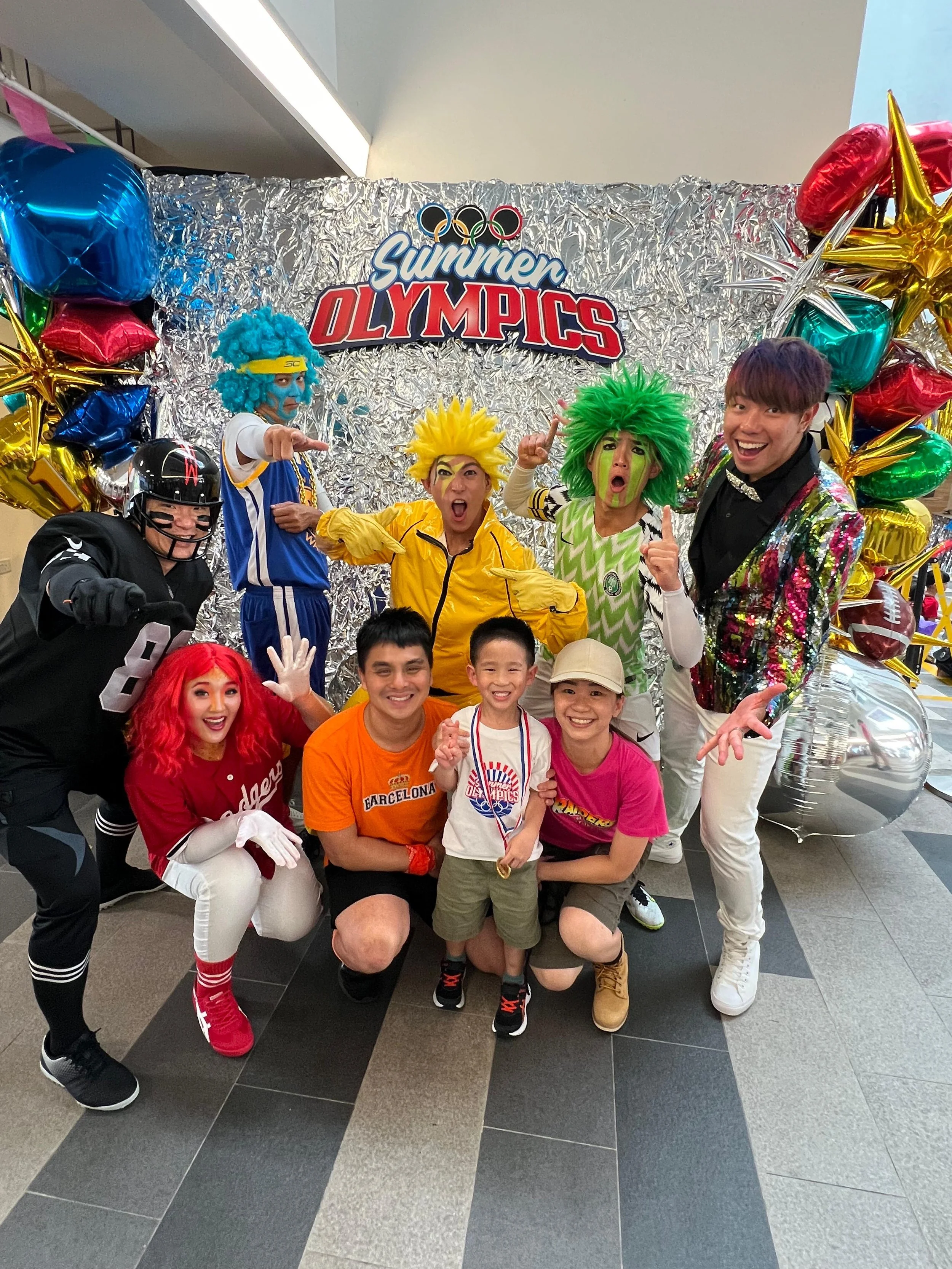 Group of five adults and four children dressed in colorful costumes, celebrating at a Summer Olympics event with balloons and a silver foil backdrop.