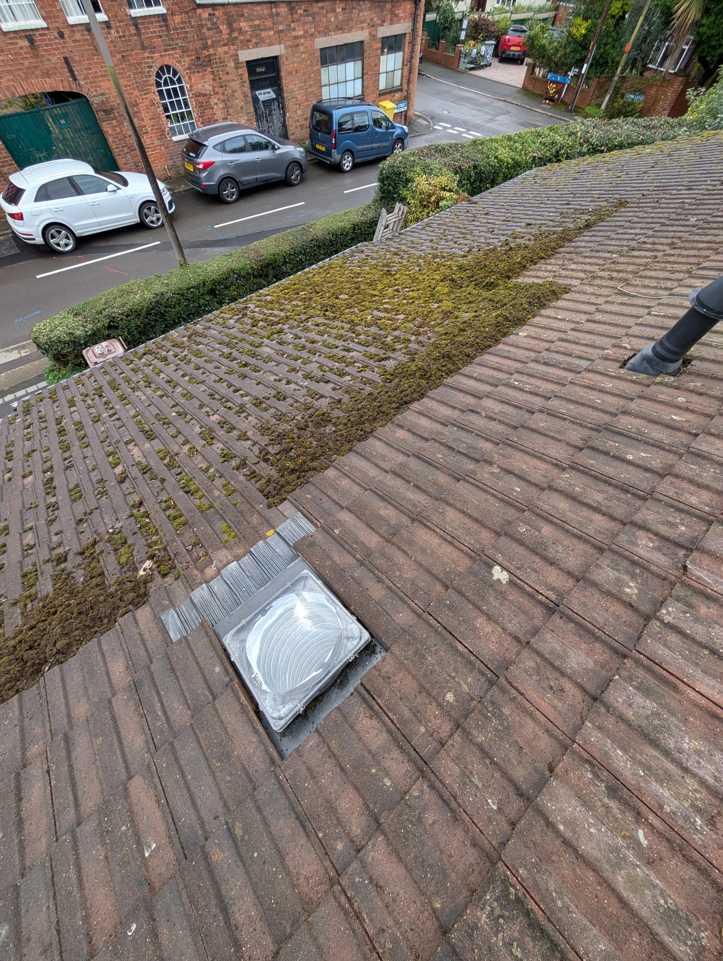 A roof with moss and algae growth, ventilation duct, and the edge of the roof with bricks. Below is a street with parked cars and a hedge, with a building with brick walls and windows.