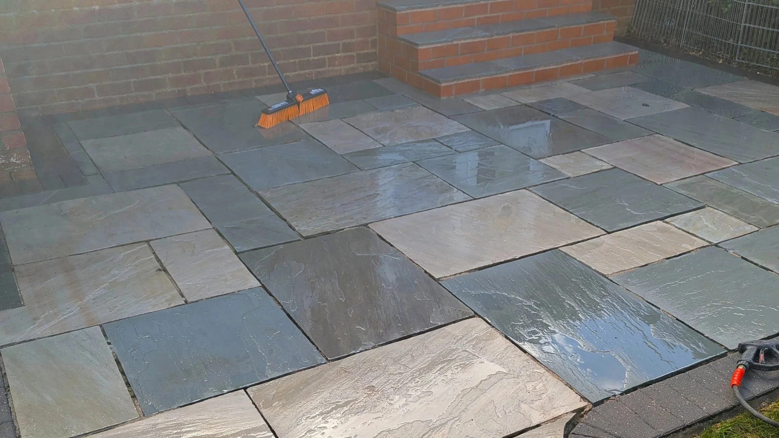 Wet stone patio outside a brick building with stairs, broom leaning against wall, and power cord on ground.