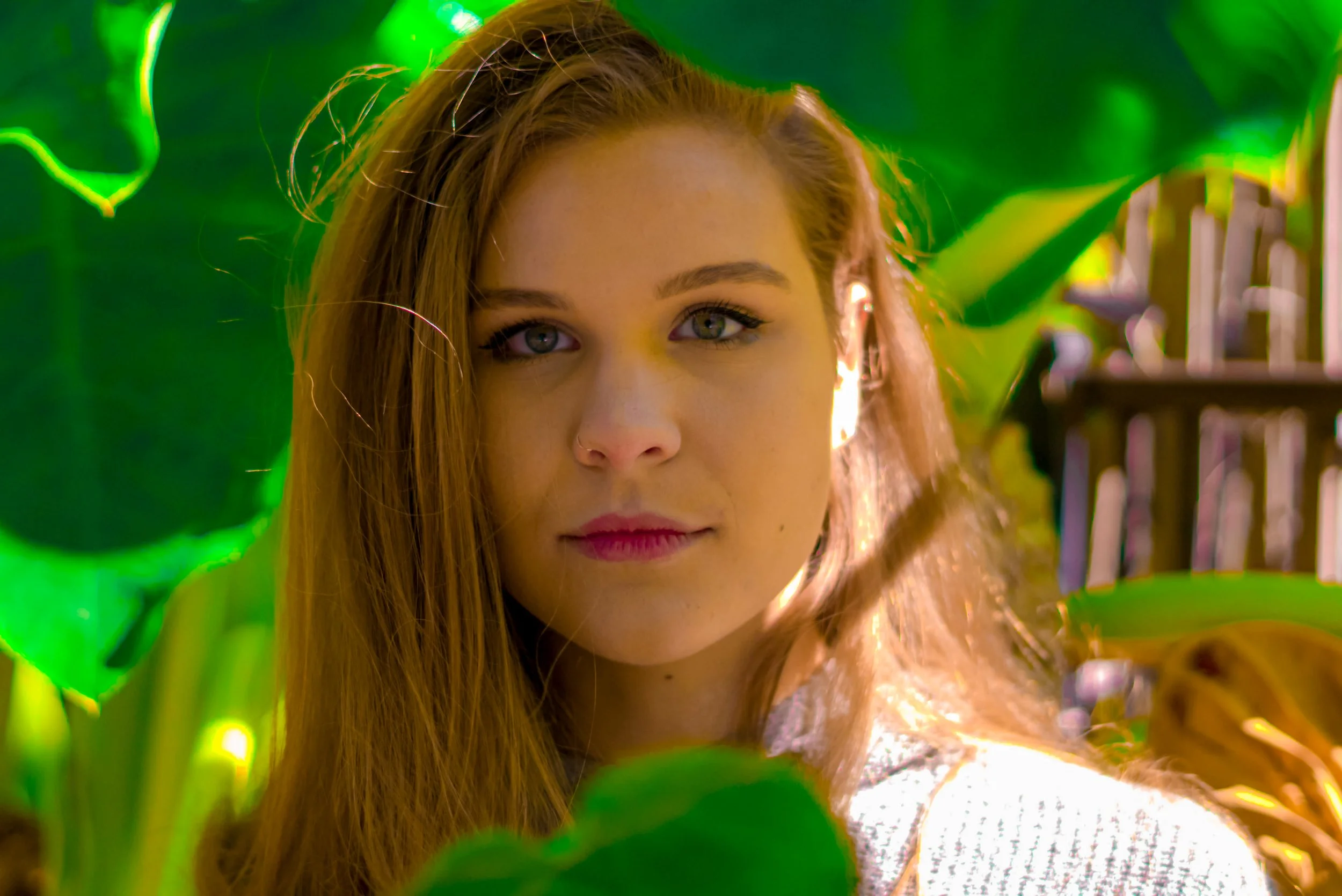 A young woman with long auburn hair and blue eyes, looking directly at the camera, surrounded by large green tropical leaves.