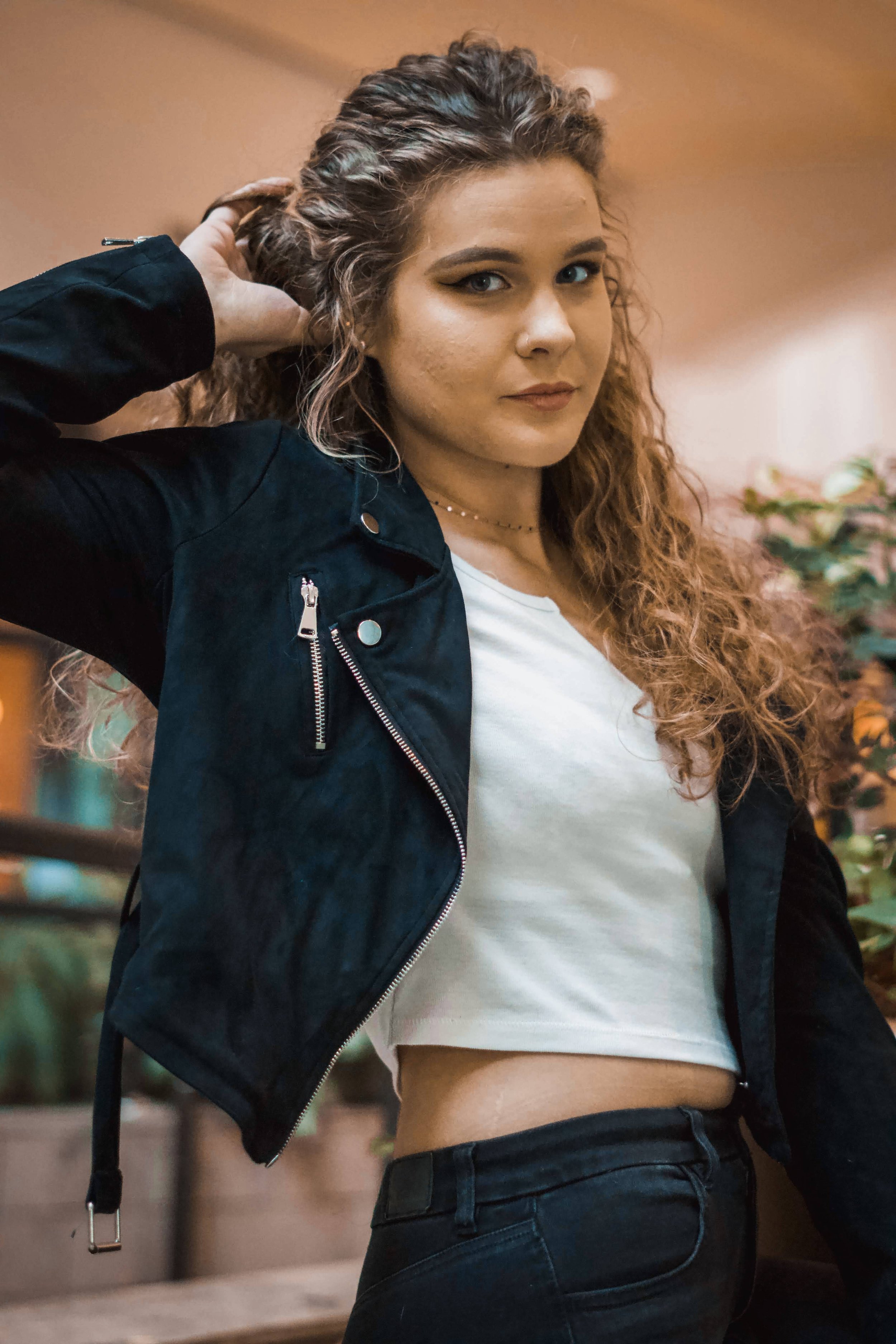 Young woman with curly hair wearing a black jacket, white crop top, and dark jeans, standing indoors with a blurred background of plants and warm lighting.