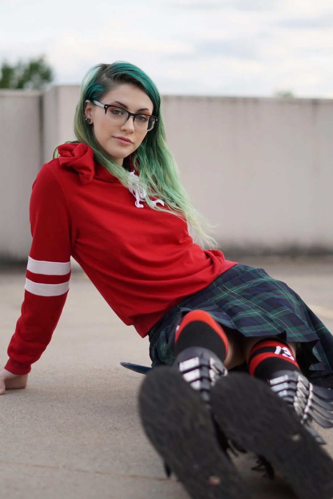 A young woman with teal and blonde hair, wearing glasses, a red hoodie with white stripes on the sleeve, plaid skirt, and knee-high socks, sits on the ground outdoors with a neutral expression.