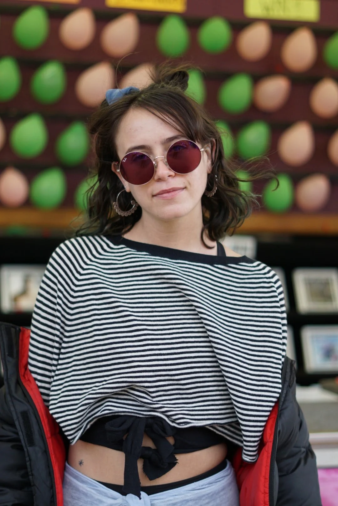 A young woman with dark wavy hair wearing red-tinted sunglasses, hoop earrings, a striped crop top, and a black jacket with red lining, standing in front of a colorful background with green and pink objects.