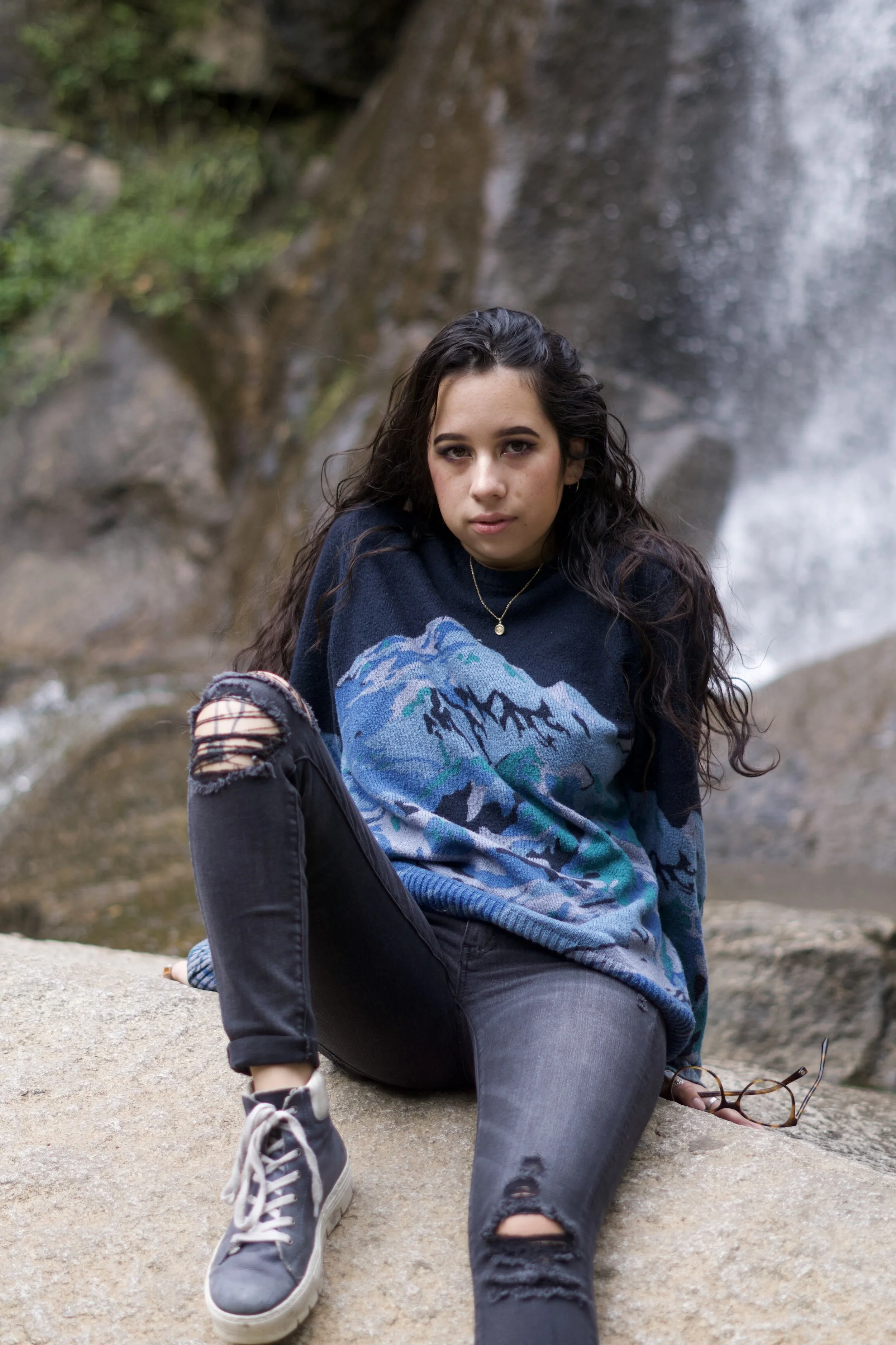 Young woman sitting on a rock outdoors near a waterfall, wearing a blue patterned sweater, ripped black jeans, and sneakers, holding glasses in her hand.