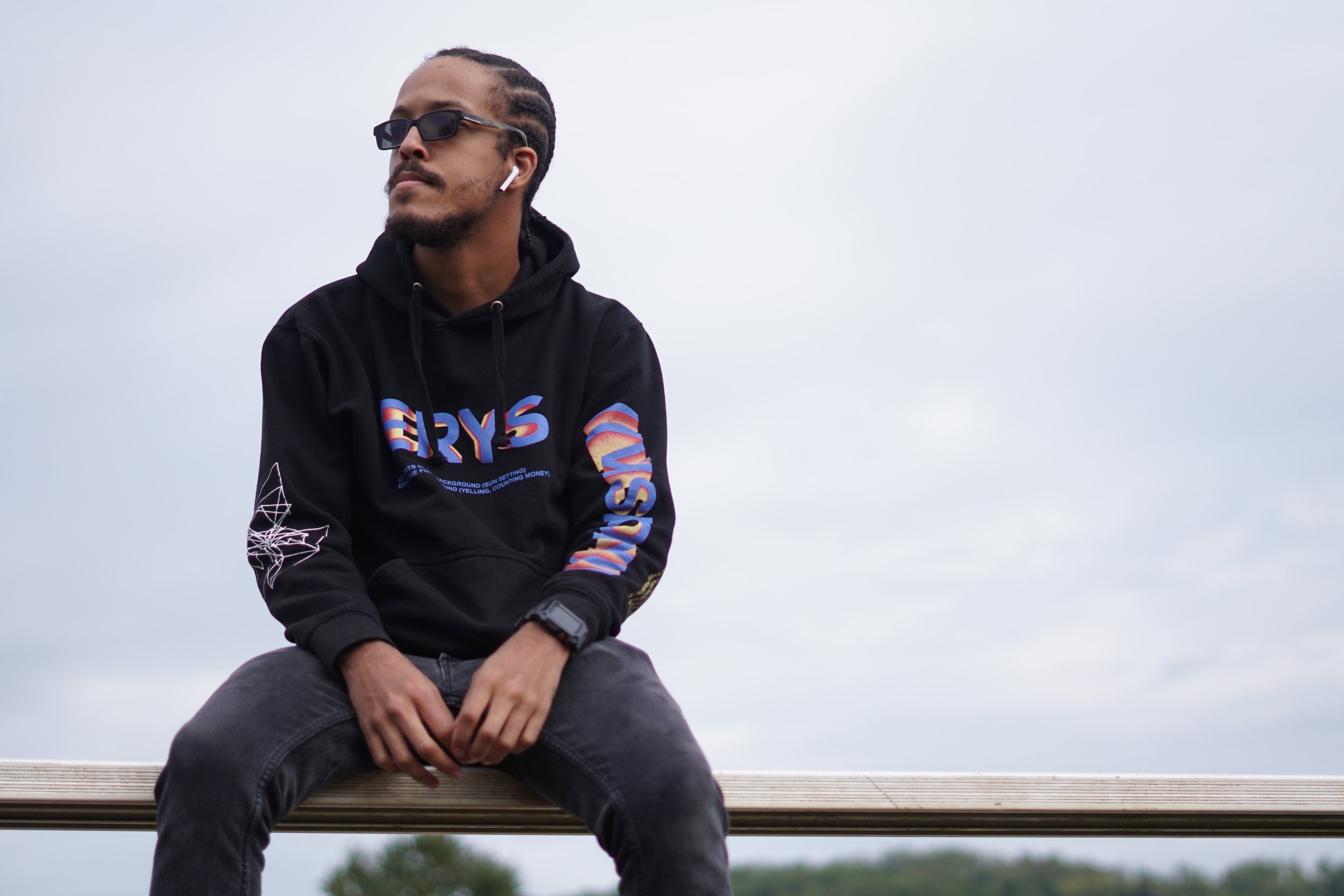 A man sitting on a wooden park bench outdoors, wearing a black hoodie with colorful text and designs, gray jeans, sunglasses, and wireless earbuds, against a cloudy sky background.