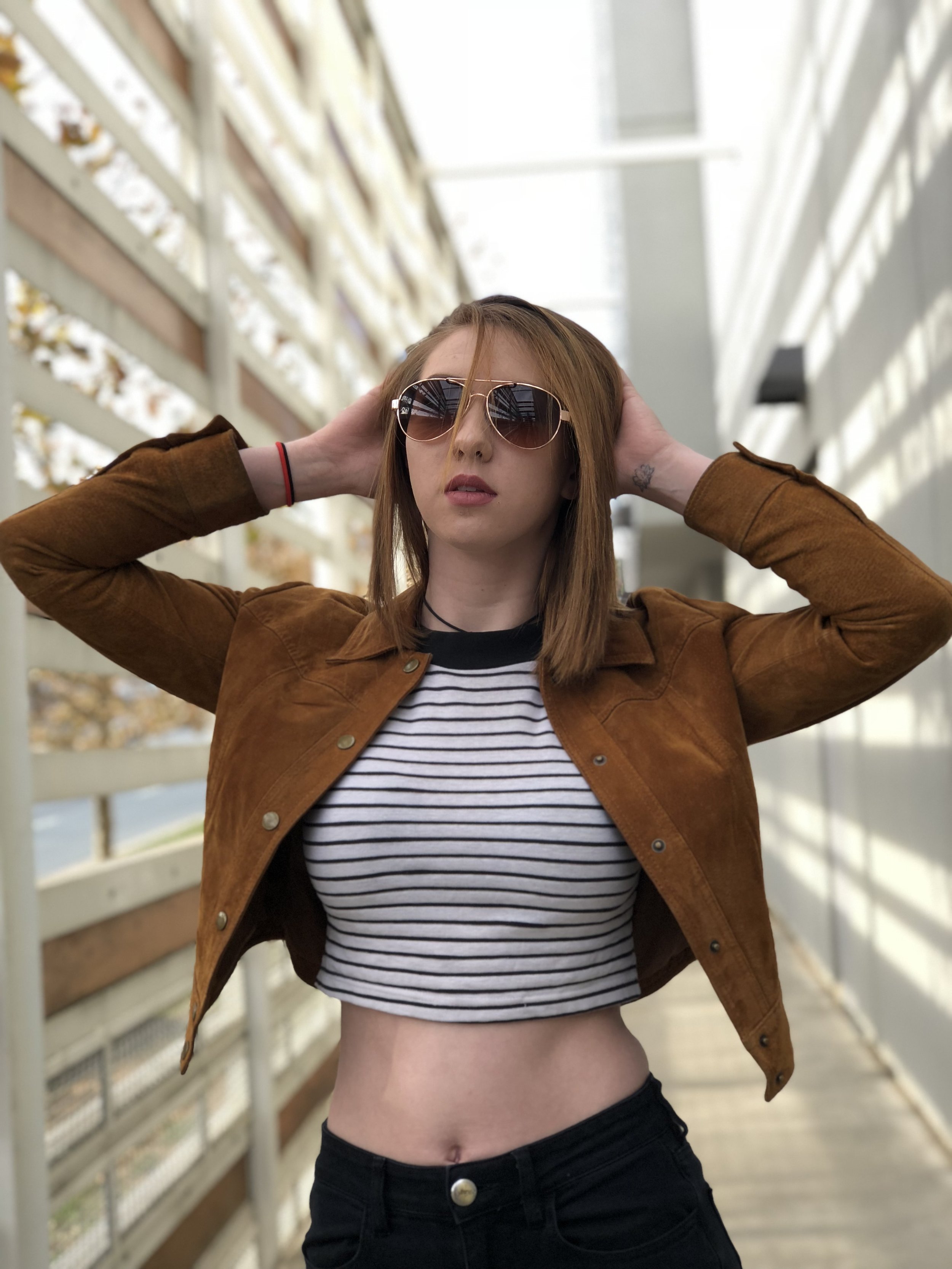 A young woman with red hair wearing sunglasses, a brown jacket, a striped crop top, and black pants standing in an outdoor corridor with a modern architectural background.