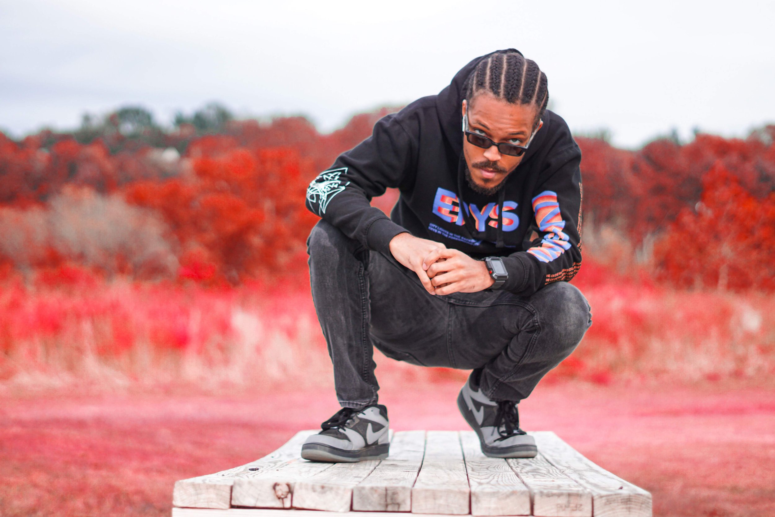 A man crouching on a wooden platform outdoors, wearing black clothing, sneakers, sunglasses, and a watch, with a background of red trees and overcast sky.