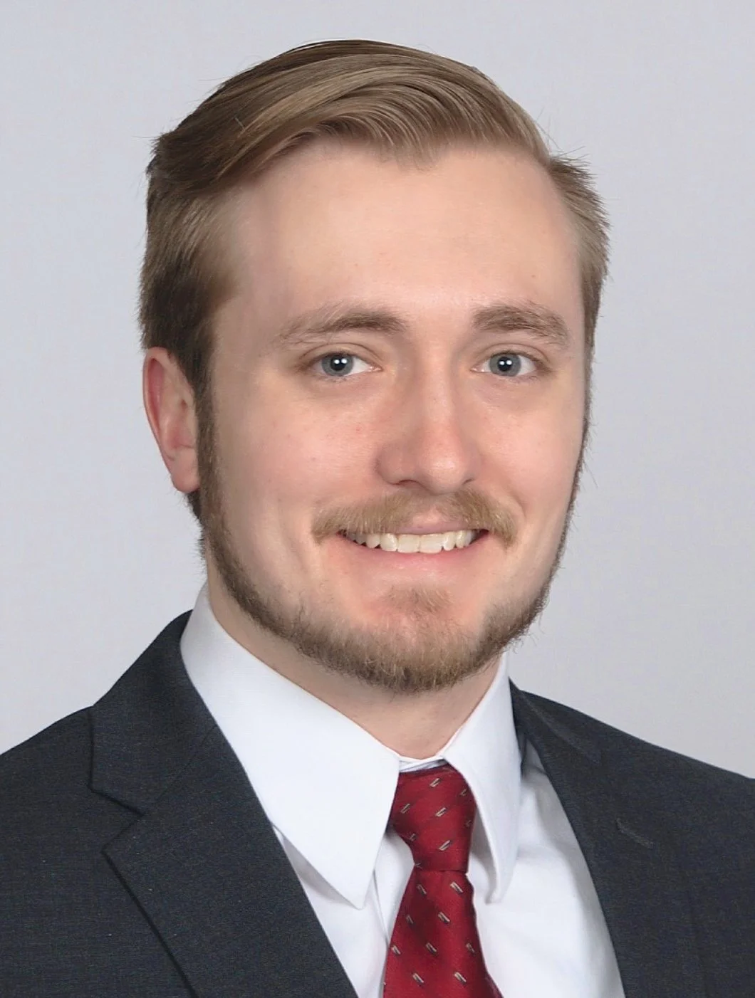 Headshot of a young man with light brown hair, a beard, blue eyes, wearing a dark suit, white shirt, and red tie, smiling.