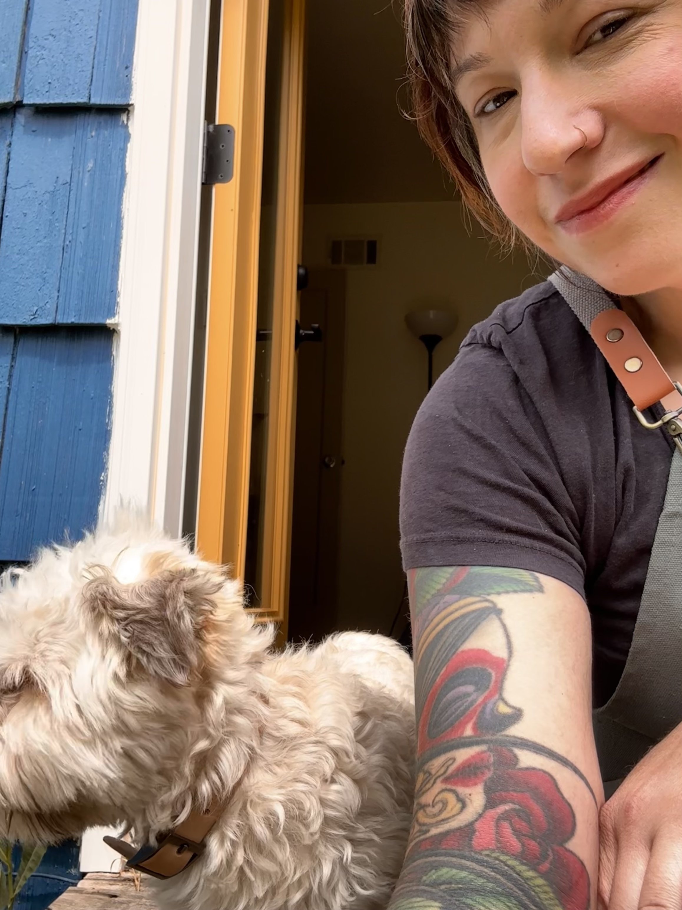 A person with short hair and a nose ring taking a selfie with a fluffy, light-colored dog wearing a collar, sitting on a porch, with part of a blue house and a yellow door frame in the background.