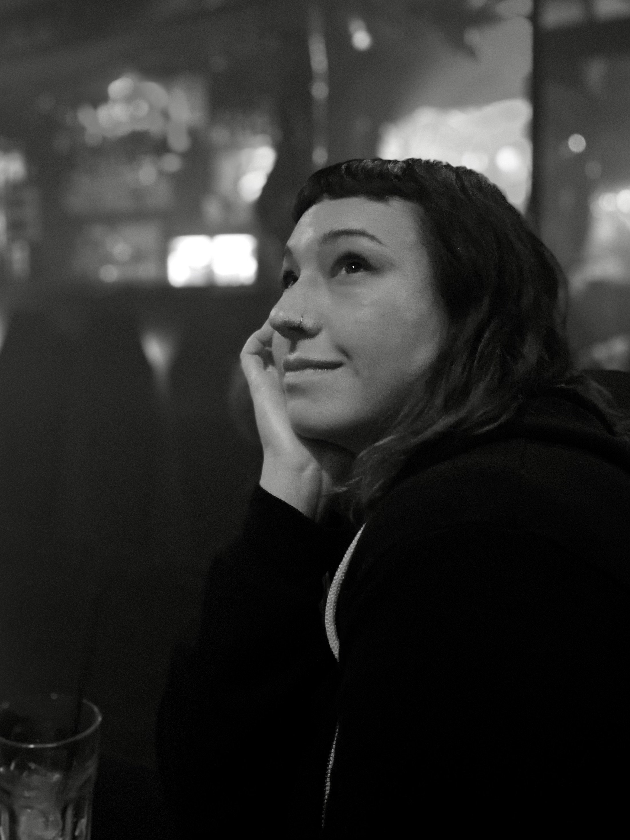 A black and white photo of a woman with dark hair, touching her face with her hand, looking upwards with a thoughtful expression, in a dimly lit setting with blurred lights in the background.