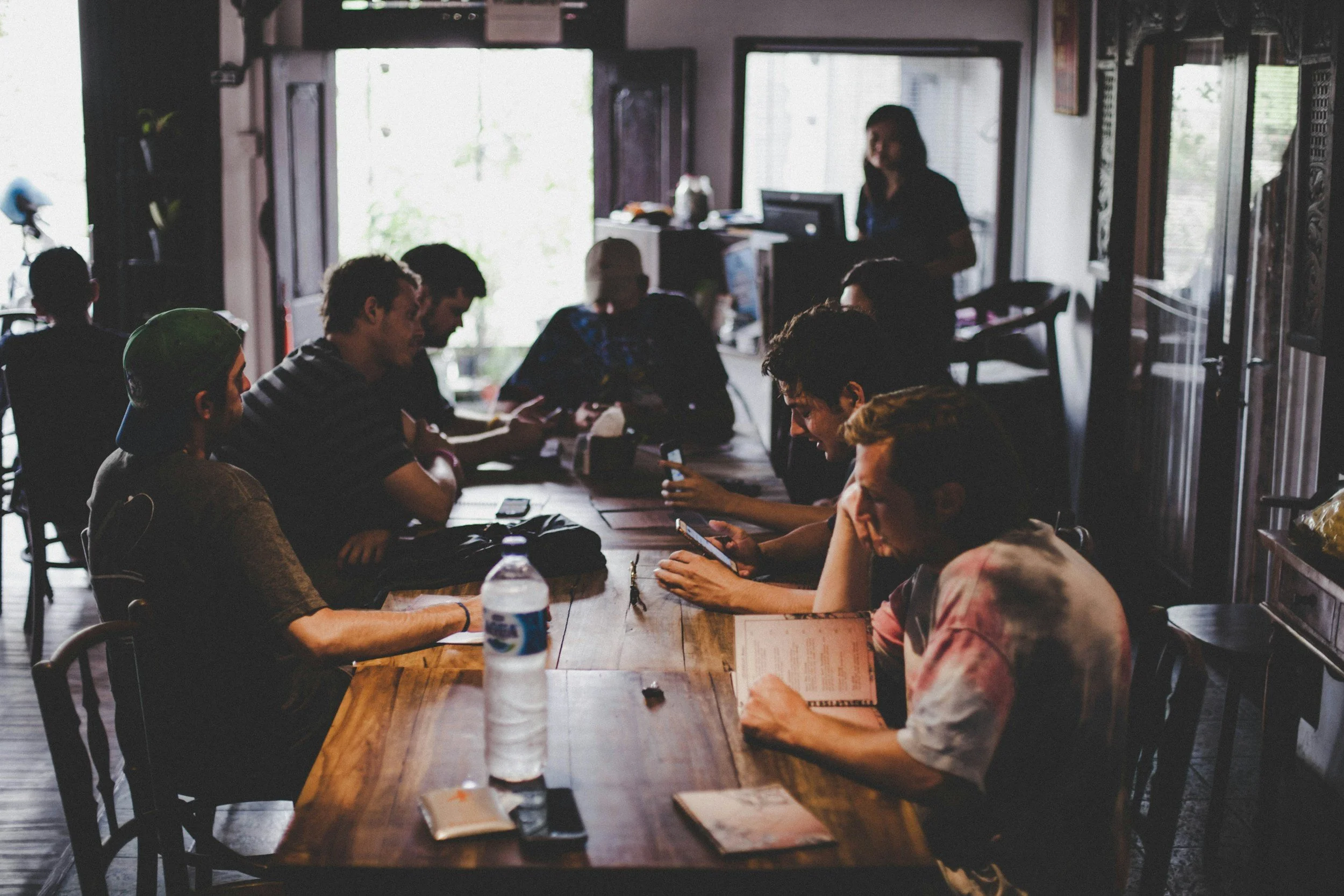 Group of young people sitting at a long wooden table in a co-working space, some looking at their phones, with a woman standing behind a counter in the background near the open door.