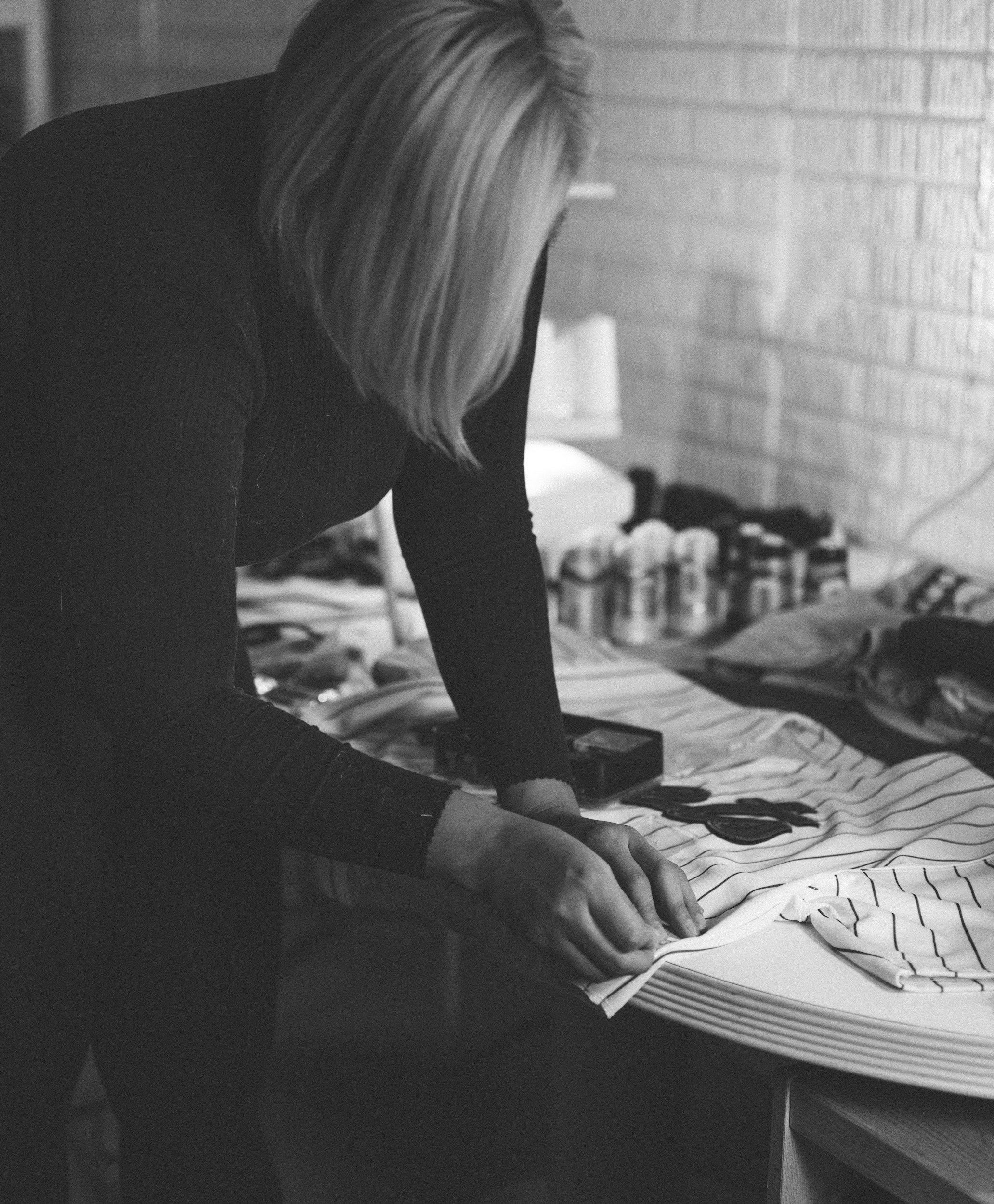A woman with shoulder-length hair is working on a fabric, possibly sewing or tailoring, on a table covered with striped cloth. The workspace has various tools and supplies, including scissors and jars, and a brick wall in the background.