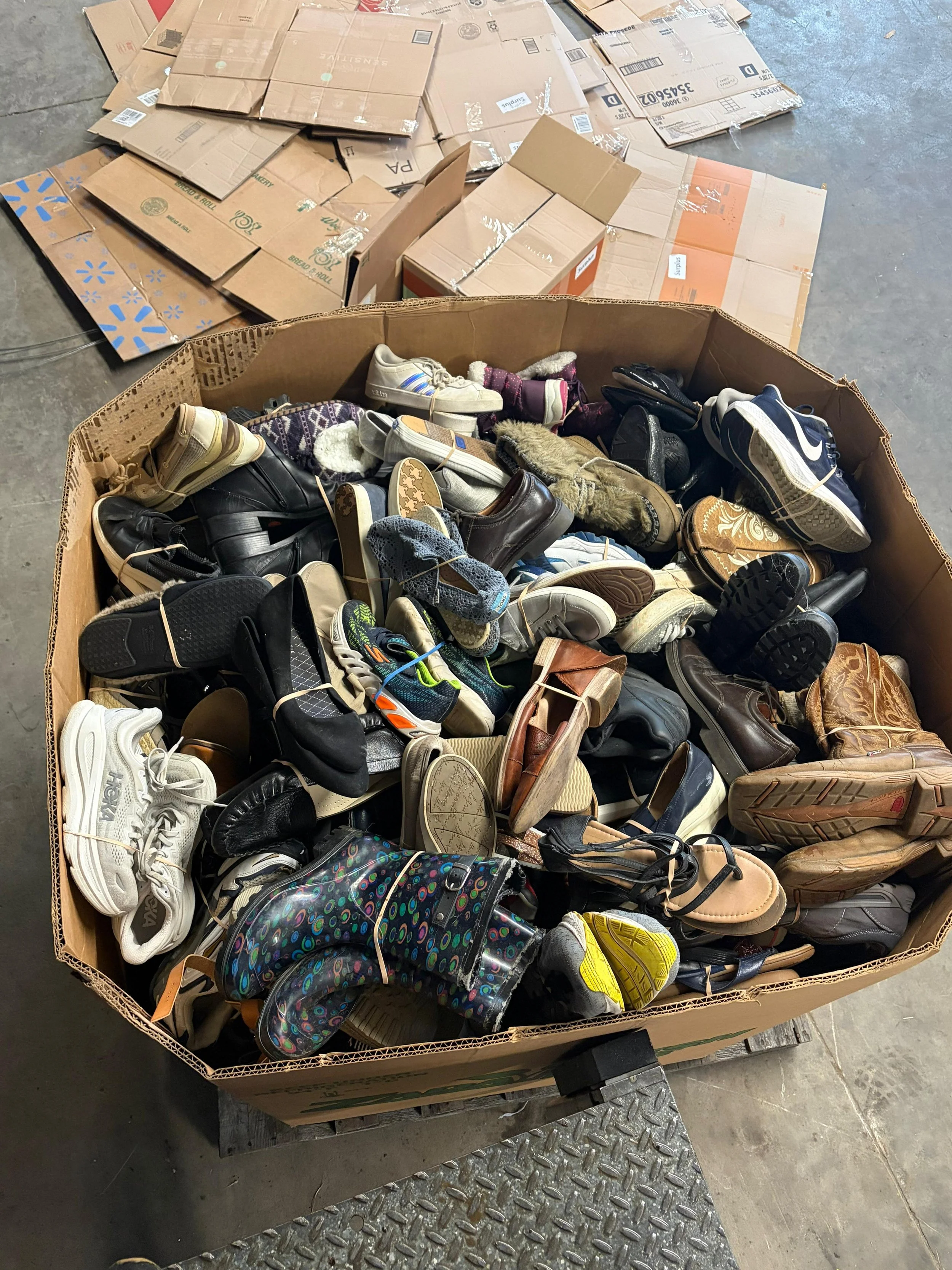 A large cardboard box filled with various shoes, including sneakers, boots, and sandals. In the background, there are flattened cardboard boxes on the floor.