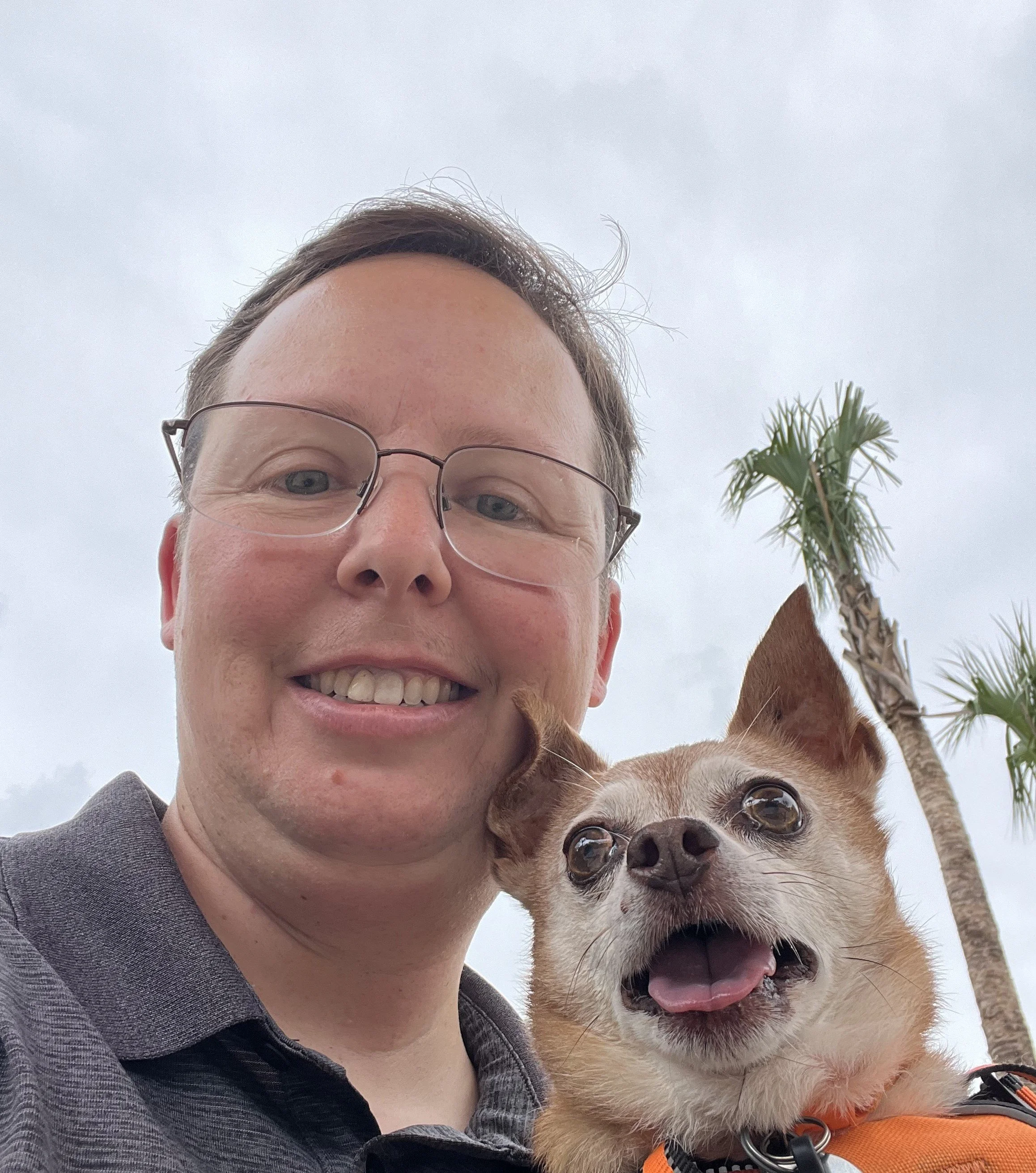 A person with glasses smiling and a small dog with an open mouth and protruding tongue outdoors, with palm trees and cloudy sky in the background.