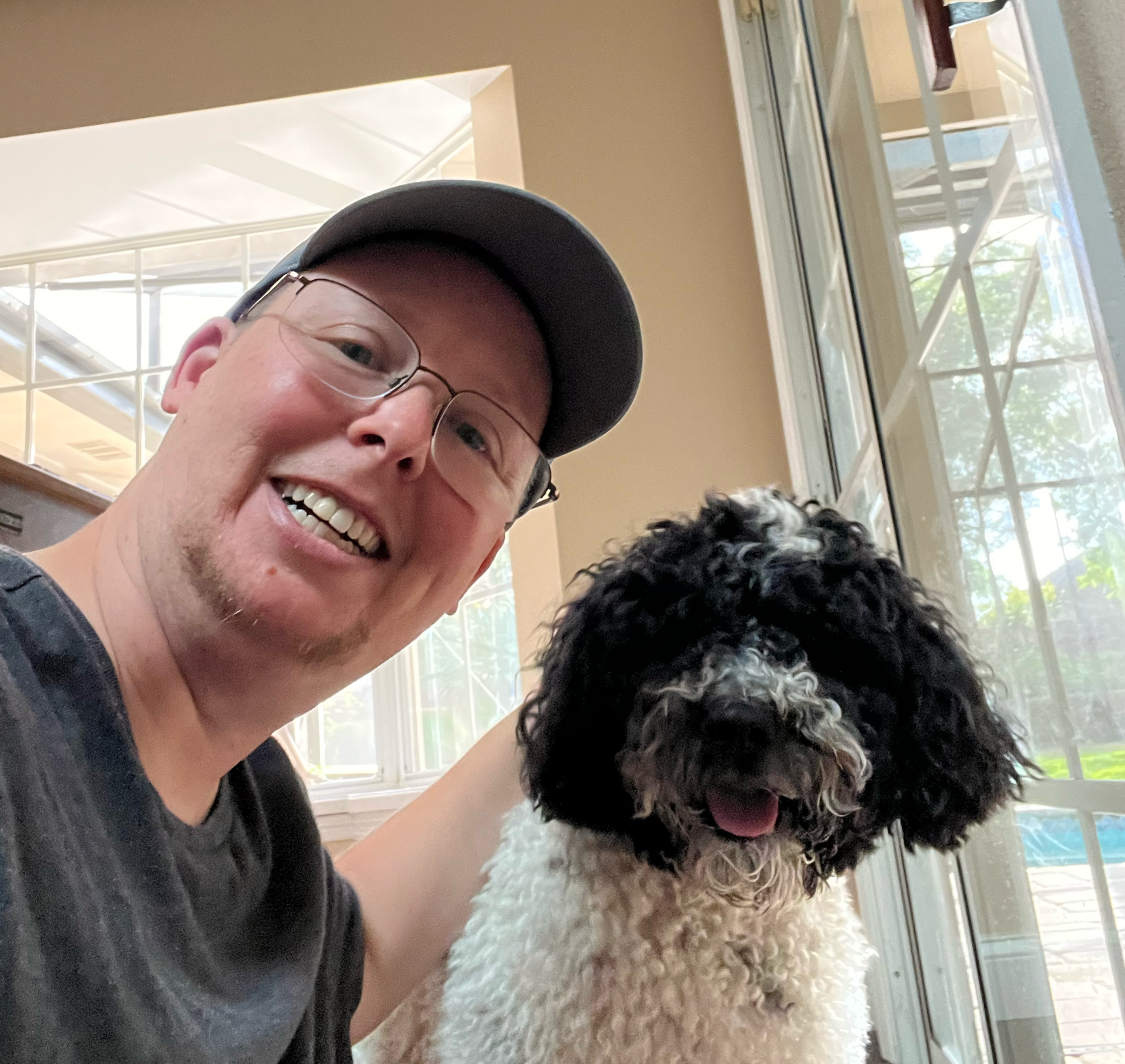 A man wearing glasses and a baseball cap smiling for a selfie with a black and white fluffy dog near a glass door with a view outside.